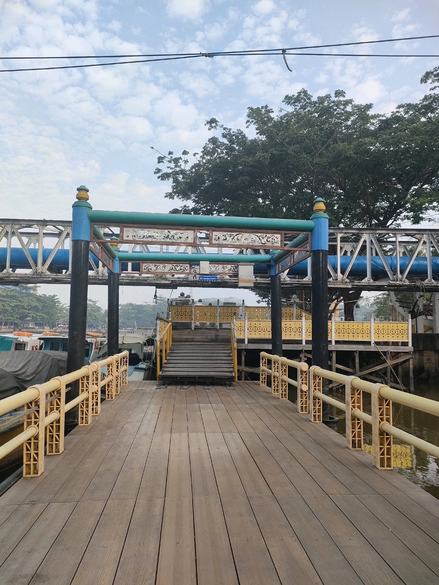 The image depicts a wooden pier leading to a covered walkway and stairs, situated in Banjarmasin, Indonesia. The pier has yellow railings. Several boats are docked alongside the pier. In the background, a large green tree and a pedestrian bridge can be seen. The sky is cloudy. The scene appears to be a public transportation hub or a recreational area by the water.