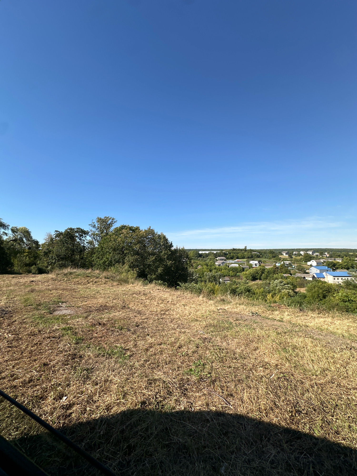 The image is a wide-angle shot of a rural landscape under a clear blue sky. In the foreground, dry, yellowed grass covers a sloping hill. Beyond the hill, a cluster of houses with blue and white roofs are visible amidst lush green trees. The settlement appears to be a village. The overall impression is of a sunny day in a peaceful, rural area. The location is identified as Makiv, Ukraine. There are no people or animals visible in the frame. The composition suggests the photo was taken from a slightly elevated position, looking down towards the village.