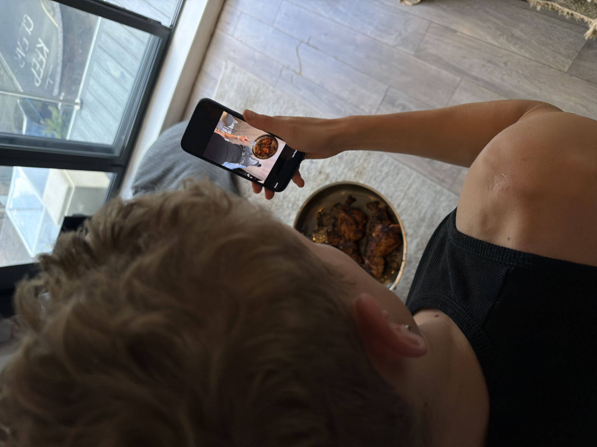 In a close-up shot, a person with short, blonde hair is holding a smartphone, capturing an image of a meal. The meal, consisting of chicken pieces in a sauce, is presented in a metallic dish on a table. The phone screen displays a reflection of the person, indicating they are taking a selfie or a picture of the dish with themselves in the frame. In the background, a large window overlooks an urban environment, with a glimpse of a "KEEP CLEAR" sign on the street, suggesting the location is in the city of London, United Kingdom. The person is wearing a black sleeveless top. The overall setting appears to be indoors, possibly a dining area or living space with a view of the city.