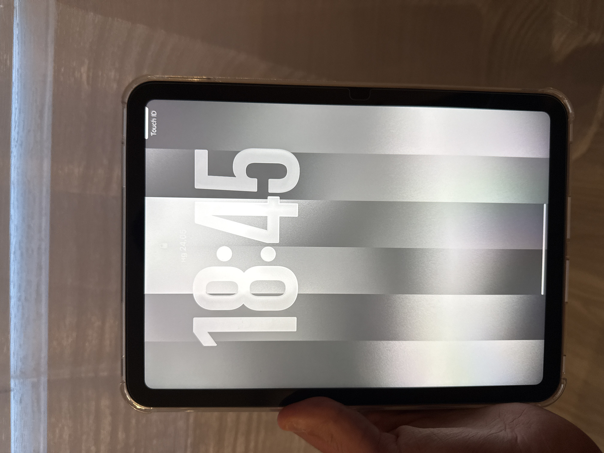 An image of a tablet displaying the time 18:45. The screen also shows "Touch ID" in the top left corner and "hg 24.05" below the time. The tablet is encased in a clear protective case. A hand is partially visible at the bottom of the frame holding the tablet. The background is blurred. The city of Lovech, Bulgaria is not identifiable in the image.