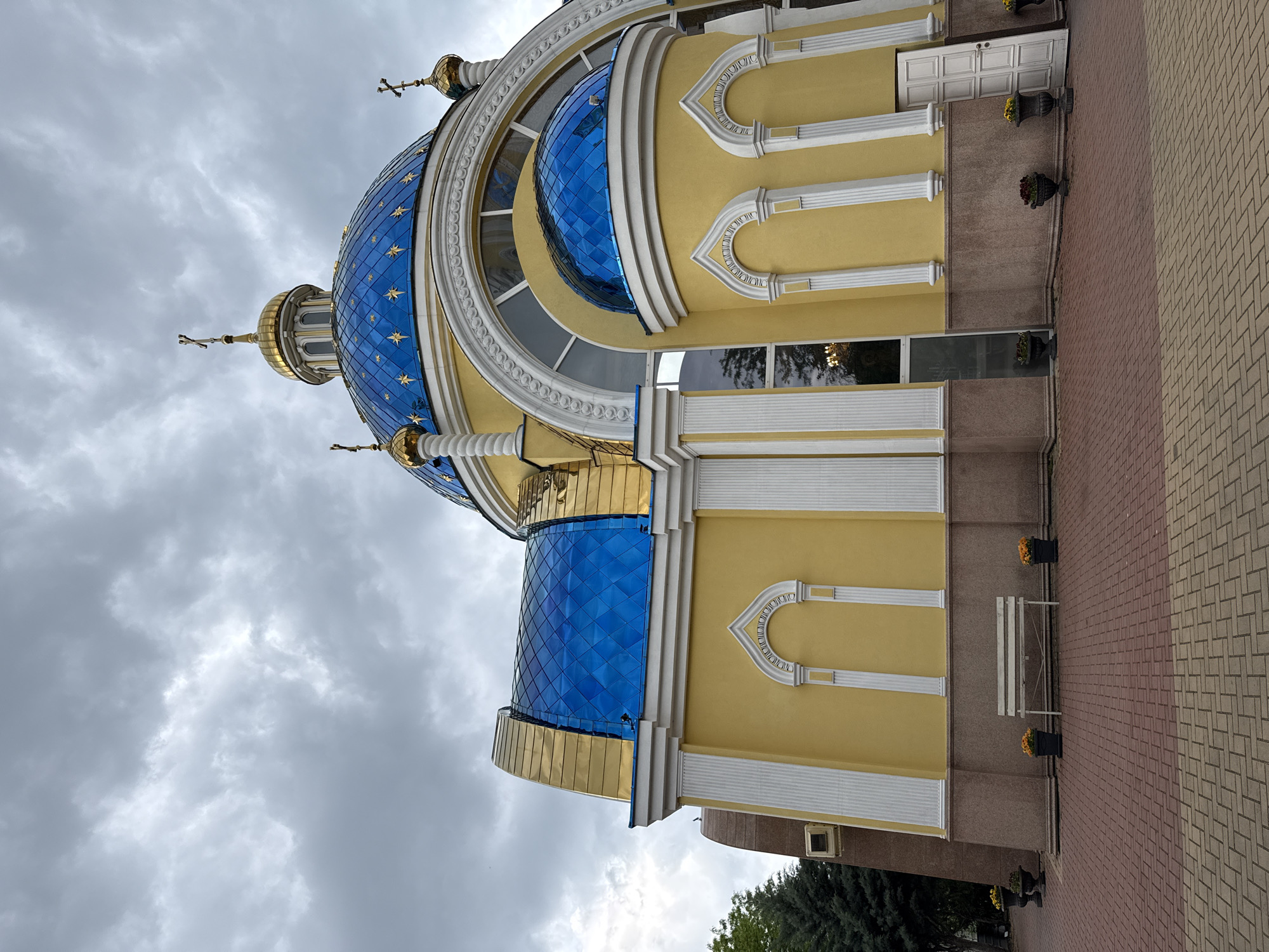 The image shows a portion of the Church of the Holy Great Martyr Marina in Plovdiv, Bulgaria. The building features a large blue tiled dome adorned with golden stars and gold accents. The walls are painted in a yellow hue with white architectural details, including arched recesses and vertical columns. Below the yellow section, there are sections of beige and brown wall with white trim. There are benches and plants in dark planters near the base of the wall. The ground is covered in a brickwork pattern. The sky is overcast with grey clouds.