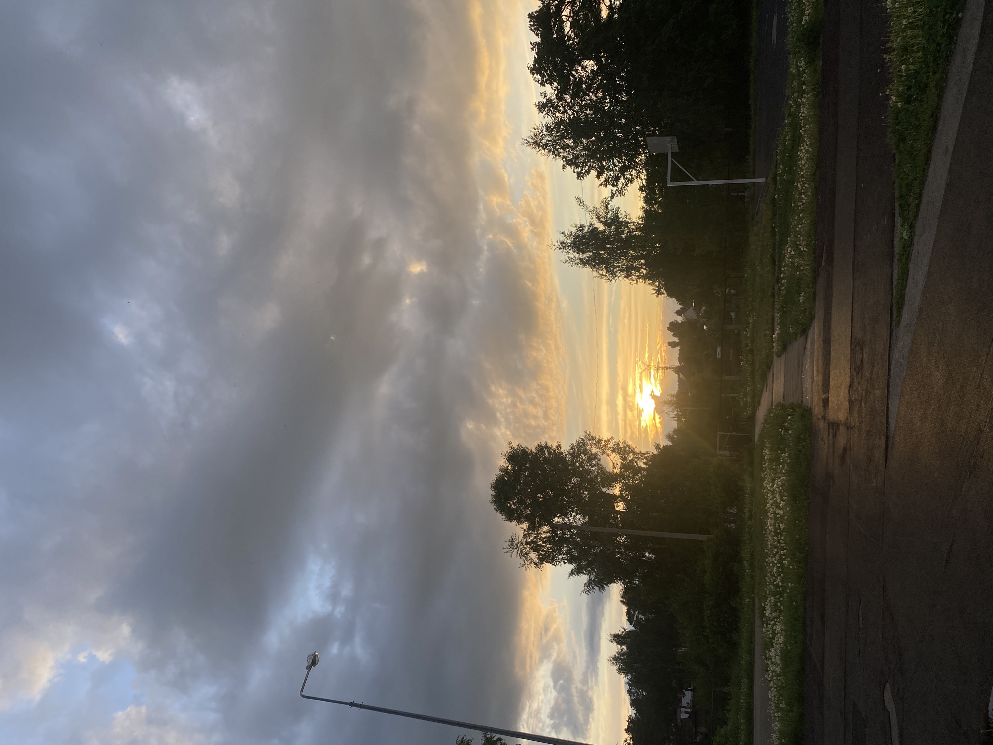 The image shows a landscape taken near dusk or dawn in what is possibly the city of Chaikovsky, Russia. In the sky, dark, overcast clouds dominate the majority of the scene, with streaks of golden light breaking through. A line of trees silhouettes against the horizon where the sun seems to be positioned, obscuring direct visibility. The foreground includes a paved area and a strip of grass, possibly a sidewalk and roadside verge. A street lamp post is visible on the left side of the image, and a basketball hoop and net are seen just beyond the trees in the middle-ground. The scene lacks any discernible human presence or direct evidence confirming its location in Chaikovsky. The image seems to capture a moment of natural beauty under a dramatic sky.