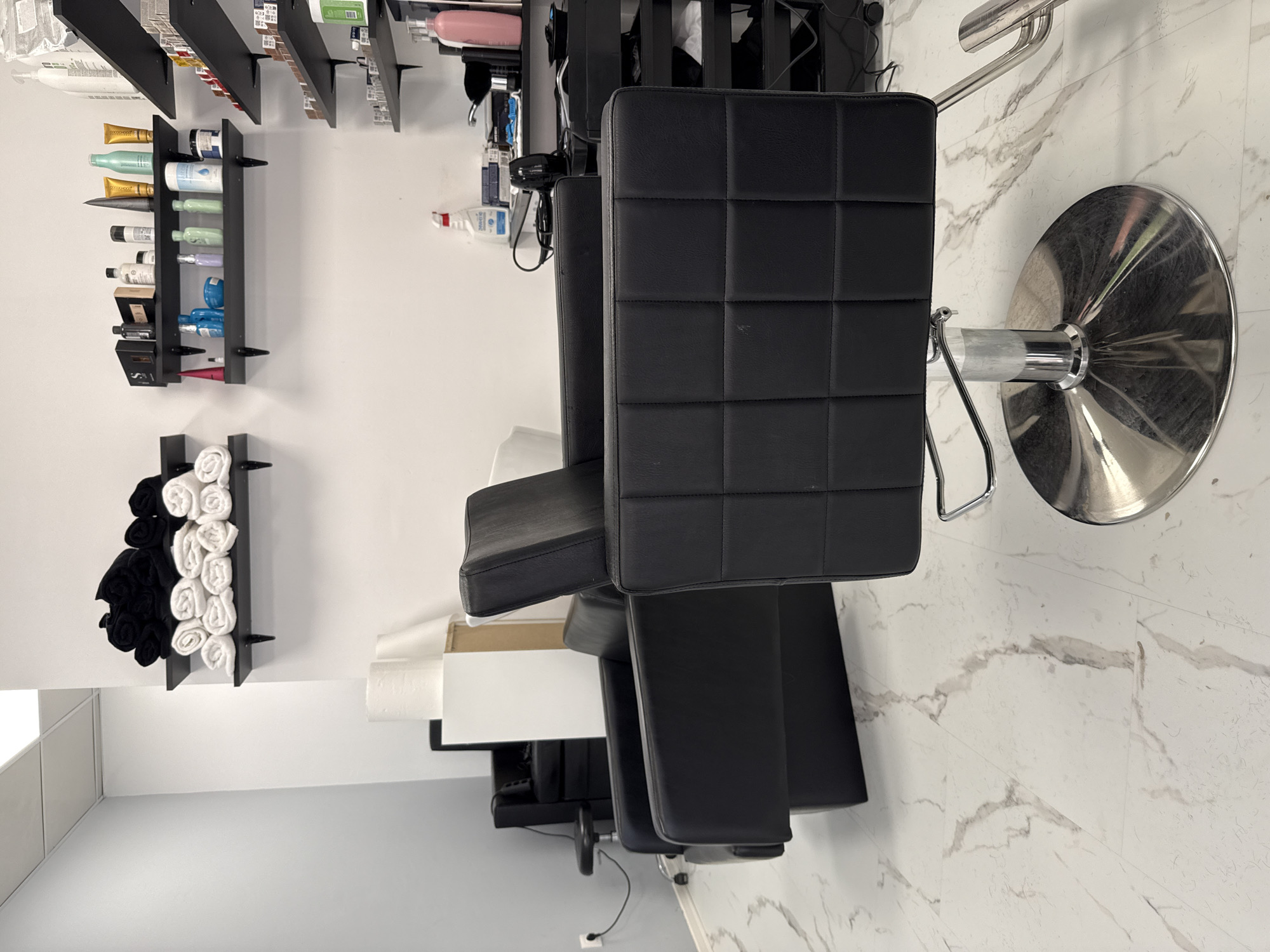 The image depicts the interior of what appears to be a hair salon. Two black, cushioned chairs with silver bases are prominently featured, suggestive of hairstyling or barber chairs. On the wall, there are shelves displaying hair care products, towels, and other salon-related items. The environment is clean, with white walls and marble-like flooring. There are no people visible in the image. The image does not provide enough information to determine its exact location in Sofia, Bulgaria, or to describe any specific actions or interactions within the scene.