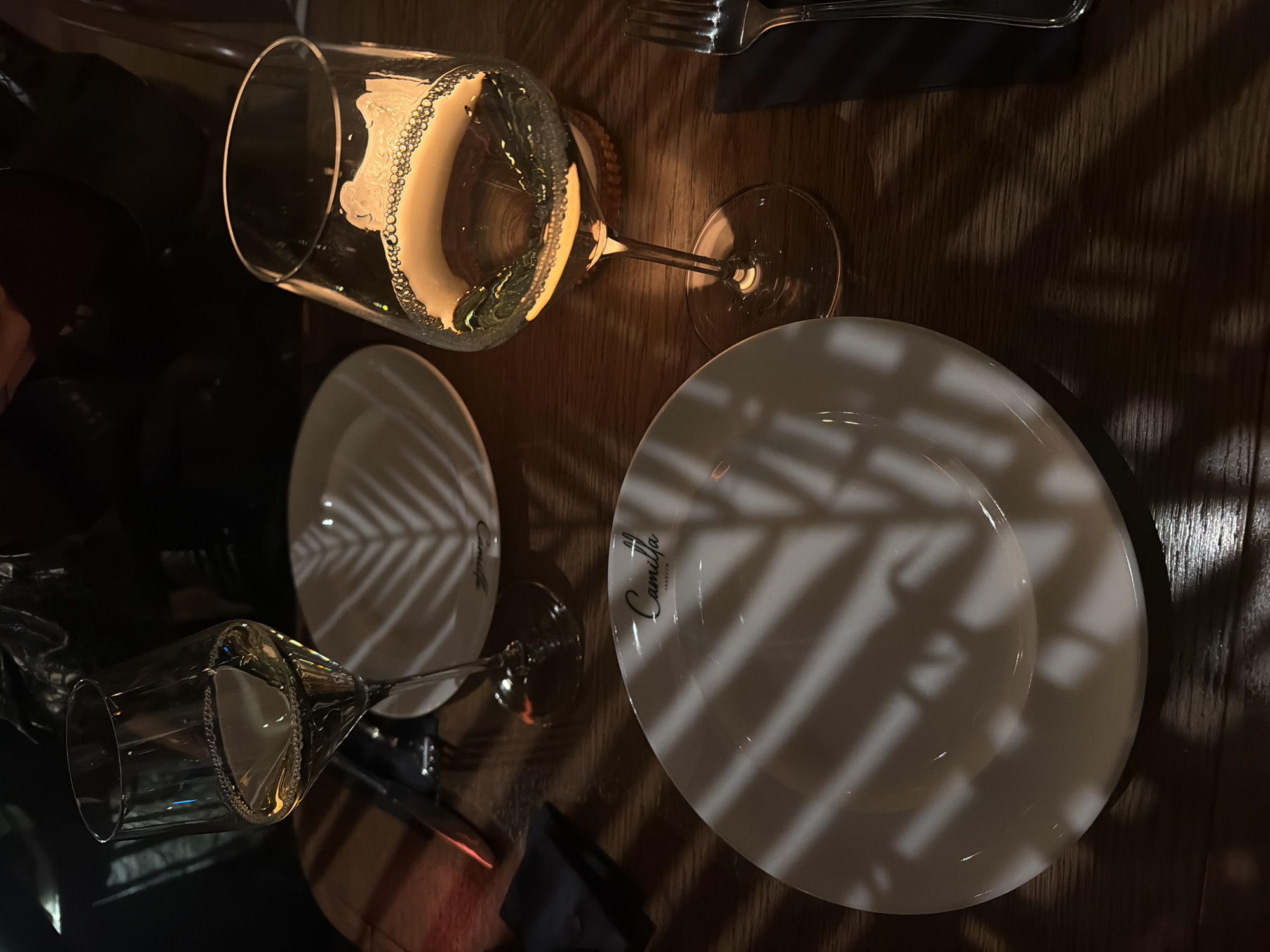The image depicts a table setting at what appears to be a restaurant. Two white plates are visible, one larger than the other, both bearing the logo "Camilla Yerevan" indicating the location is in Yerevan, Armenia. There are two wine glasses, one containing a bubbly beverage. Cutlery is present, along with a dark napkin. Shadow patterns fall across the plates and table, suggesting light filtering through foliage. The setting is indoors, with a dimly lit, refined atmosphere. No people are directly visible, but the arrangement of the place settings indicates an anticipation of diners.