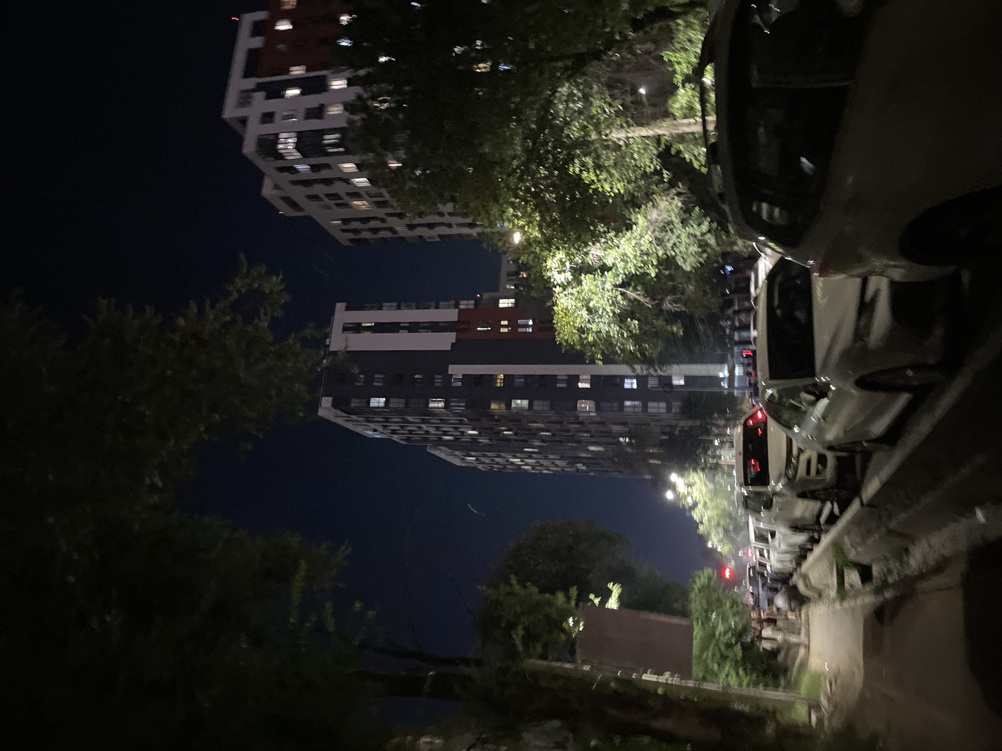 The image depicts a nighttime scene on a city street in Almaty, Kazakhstan. A row of parked cars lines the right side of the road, with a sidewalk and trees on the left. Several tall apartment buildings are visible in the background, with lights on in some of the windows. The sky is dark, indicating it is late at night. A few people are walking on the sidewalk. The street appears to be busy, with traffic lights and streetlights visible. The overall atmosphere is one of urban night life.