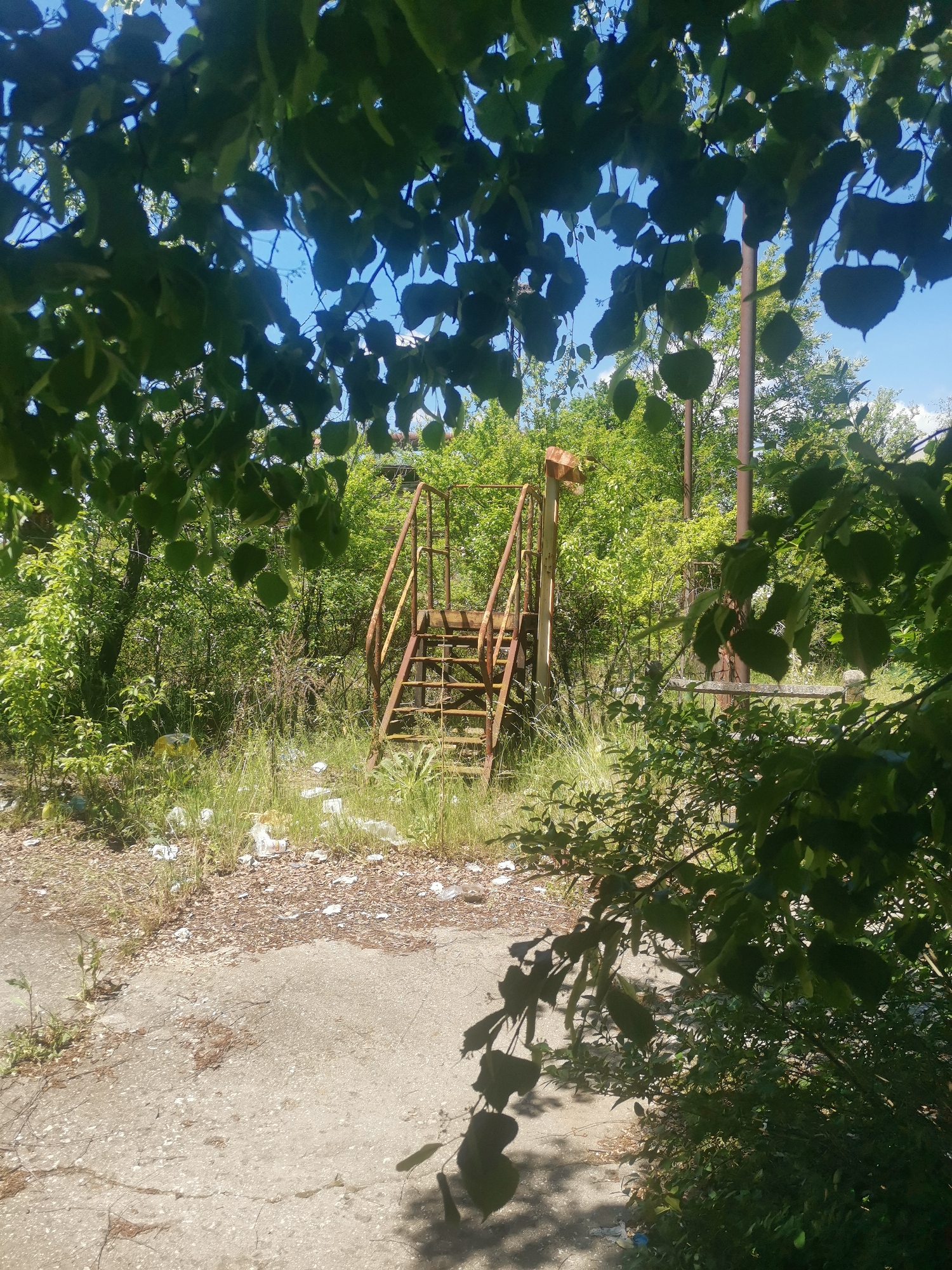 The image shows a dilapidated outdoor scene in what appears to be a neglected area. In the center of the frame, partially obscured by overgrown vegetation, is a rusty metal staircase, indicating a structure or platform that has been abandoned. The ground around the staircase is covered in debris, including what appears to be litter and fallen leaves. The foreground and background are dominated by lush greenery, with trees and bushes partially obscuring the view. A concrete slab is visible in the lower part of the image, suggesting a paved area that has fallen into disrepair. Two metal poles can be seen behind the staircase. No people or animals are visible in the frame. The location is Verinsko, Bulgaria.