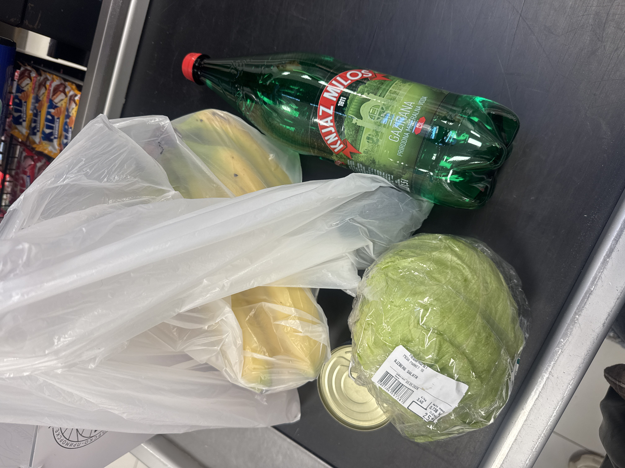 On a checkout conveyor belt, groceries consisting of a plastic bag with bananas, a can, a head of lettuce in plastic wrap, and a bottle of "Knjaz Miloš Gazirana" mineral water. In the background are "Kidy" chocolate bars. The lettuce's label indicates "Mega Market 10" and "AJZBERG SALATA," suggesting the scene is in a MEGA MARKET location in Budva, Montenegro. The "Pakovano" date on the label is 05.05.2026. The label indicates the mass is 0,738 kg and the price is 2.5 euro. The "Knjaz Miloš Gazirana" mineral water is in a green 1.5L bottle.
