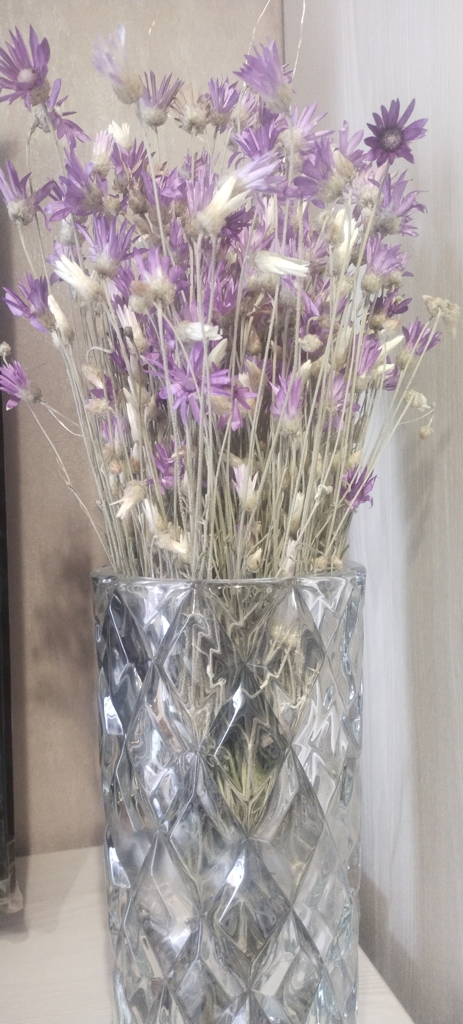 A close-up, eye-level shot shows a clear glass vase filled with dried purple and white flowers. The vase has a diamond-shaped, textured pattern. The background consists of a light brown wall and a light-colored curtain on the right. The image appears to be taken indoors, likely in a residential setting. There is no indication of a specific location in Pisochyn, Ukraine, or any other city.