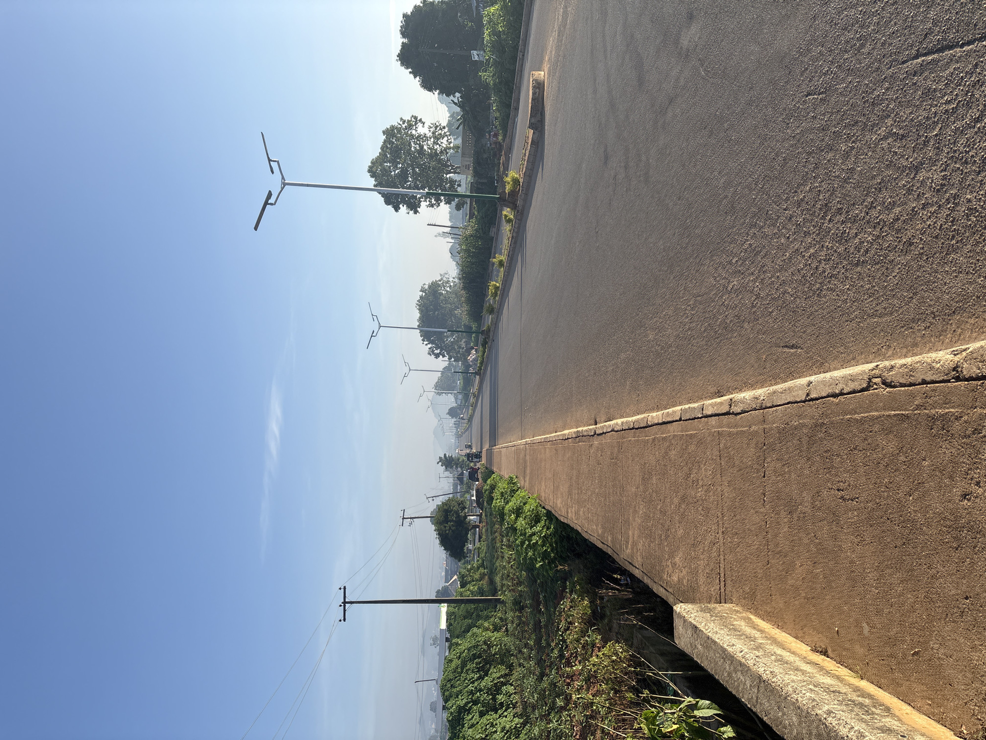 The image depicts an empty paved road in Bukuru, Nigeria, on a clear day. The road is flanked by a concrete sidewalk and a grassy verge with vegetation. Several streetlights are visible along the roadside, with one prominent streetlight in the foreground. Power lines and utility poles are also present, stretching across the scene. In the distance, a few vehicles, possibly tricycles, are moving along the road. The overall environment suggests a moderately developed urban or peri-urban area, with a clear sky and the presence of greenery.