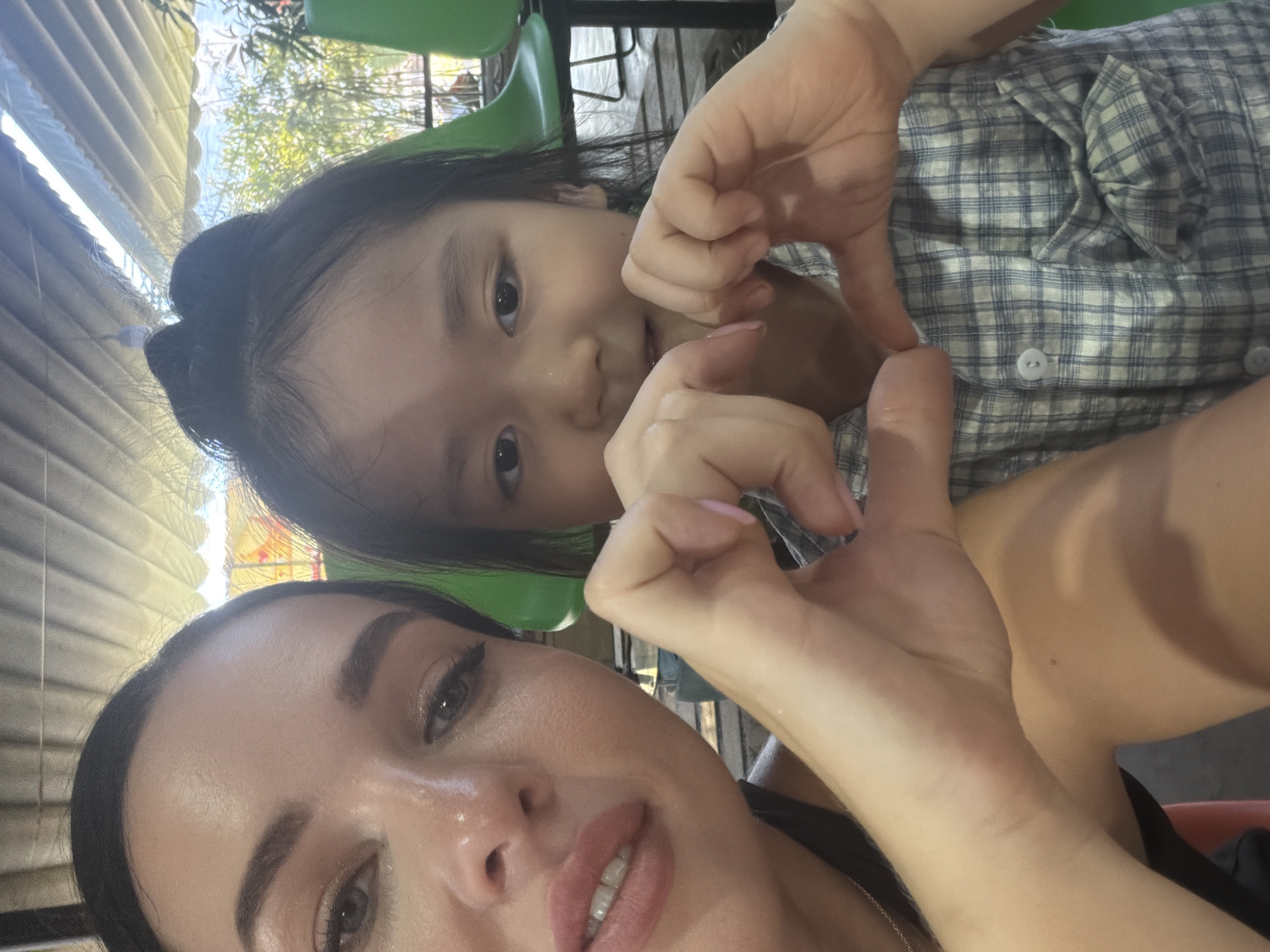 A woman and a child are posing for a selfie. They are both making a heart shape with their hands. The child is wearing a plaid shirt. The woman is wearing a black top. The background appears to be an outdoor seating area with green chairs and an awning. The location is Nam Nha Trang Ward, Vietnam.