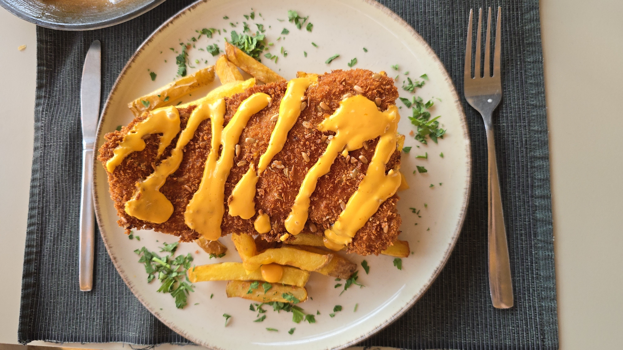 A close-up, overhead shot shows a plated meal consisting of breaded chicken or fish, served with french fries and drizzled with a bright yellow sauce. Chopped parsley is sprinkled over the dish as a garnish. The plate is positioned on a dark, textured placemat. A fork rests below the plate, and a knife is partially visible above it. The background is blurred, suggesting an indoor setting. The location is a restaurant in Staro Myasto, Bulgaria.