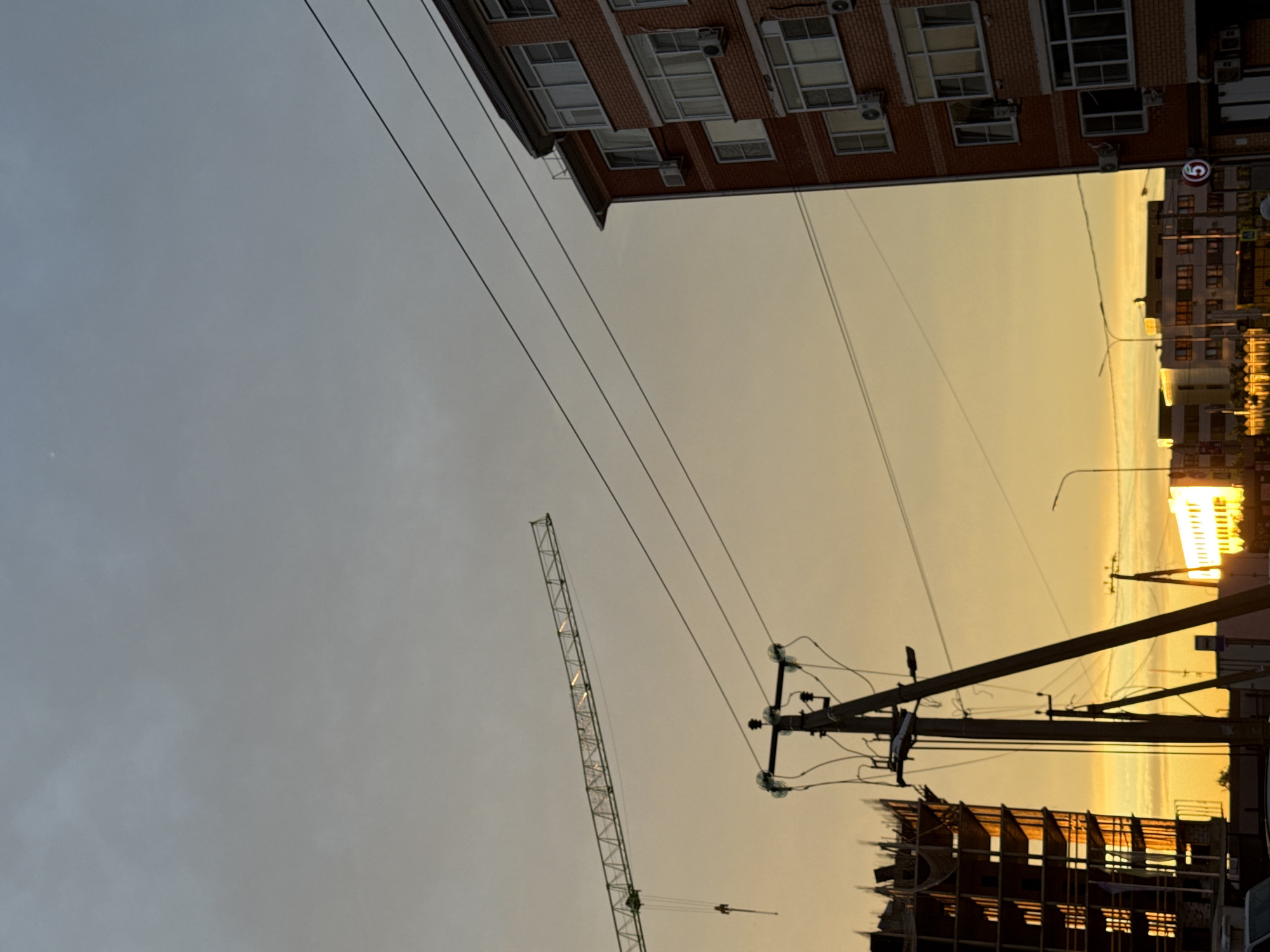 An urban scene is captured during either sunrise or sunset, evidenced by the distinct golden light permeating the atmosphere. The sky, which dominates the upper portion of the frame, transitions from a muted grey-blue on the left to a warm, vibrant golden-yellow on the right.
On the left side of the image, a multi-story red brick building with numerous windows and air conditioning units is partially visible. Several power lines extend diagonally across the sky from this side. A tall, skeletal construction crane stands in the mid-left, its metal structure reflecting streaks of the golden light.
On the right, a prominent utility pole with multiple power lines and electrical equipment stands. Further to the right and lower in the frame, a building under construction reveals its rebar and concrete framework, brightly illuminated by the golden sun. Below this, a street or urban area stretches into the distance, with other buildings and sources of light that appear to be streetlights or reflections. A red circular sign with a white number '5' is visible on the far right, possibly a traffic sign.
The low sun casts intense golden reflections on the crane, the construction site, and distant windows, creating a striking contrast against the varying sky tones. No people are visible. The scene depicts an active urban environment at a transitional time of day, highlighting ongoing development.
