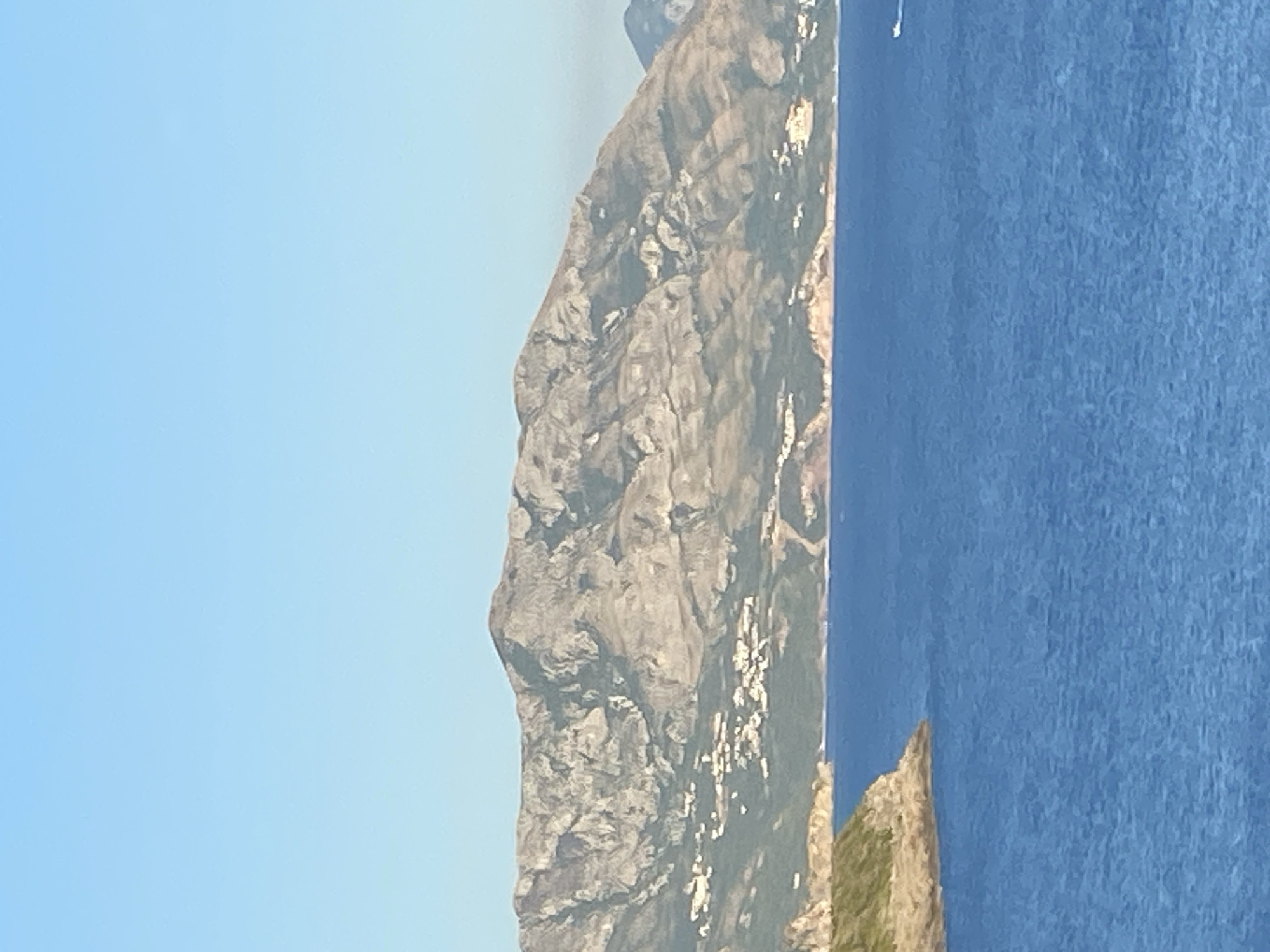 The image depicts a scenic coastal landscape, likely in Dubraljevina, Montenegro. A large, rocky mountain range dominates the background, with scattered white structures suggesting a small village or settlements nestled on its slopes. The foreground is occupied by a vast expanse of deep blue sea. A small, rocky, and vegetated island juts out into the water on the left side of the frame. In the far distance on the right, a small white boat is visible on the sea. The sky is a clear, pale blue, indicating fair weather. The overall scene is tranquil and natural, highlighting the rugged mountainous terrain meeting the calm sea.