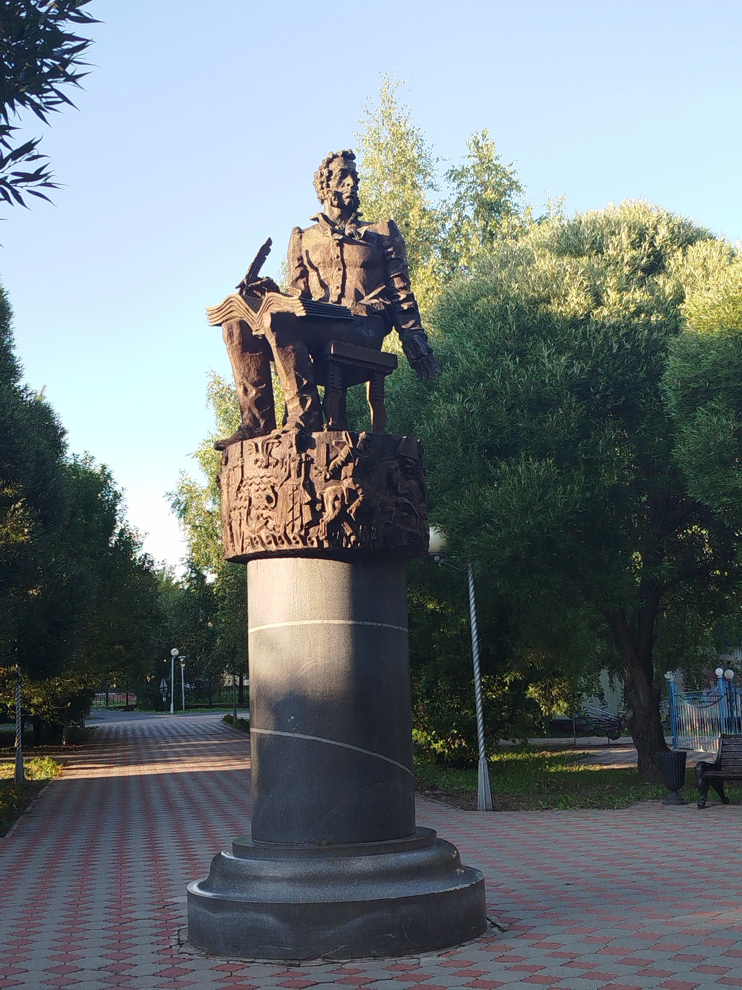 A bronze statue of a man is positioned on a stone pedestal in a park. The man is depicted seated and appears to be writing in an open book, holding a quill pen. The base of the pedestal features intricate carvings. The statue is surrounded by lush green trees and a pathway made of patterned red and gray bricks. The pathway leads into the distance, with streetlights visible along its sides. In the background, there is a park bench and a blue metal fence. The sky is clear and blue, indicating a sunny day. The overall scene suggests a public space in Yalutorovsk, Russia, likely a memorial or a tribute to a notable figure.