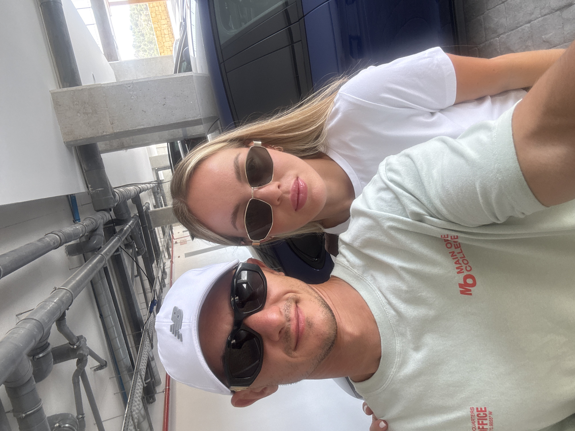 A medium shot shows a man and a woman taking a selfie in what appears to be an outdoor parking garage. The man, on the left, is wearing a white baseball cap with a gray New Balance logo, black sunglasses, and a light green long-sleeved t-shirt with red text that reads "OFFICE" and "MAIN OYE COLLEFE". He has a light stubble and is smiling faintly. The woman, on the right, is wearing a white t-shirt and gold-rimmed sunglasses. She has blonde hair and is looking towards the camera. A dark blue car is visible behind them. Above them, a complex network of pipes and conduits runs along the ceiling. The background also shows concrete pillars and a glimpse of a hilly landscape with some trees and a stone wall. The lighting suggests it is daytime, possibly with natural light filtering in. The flooring appears to be made of interlocking tiles. The presence of the car and the architectural elements suggest an urban or semi-urban environment, potentially a residential or commercial parking area. Given the context, this location is likely within the city of Finestrat, Spain.