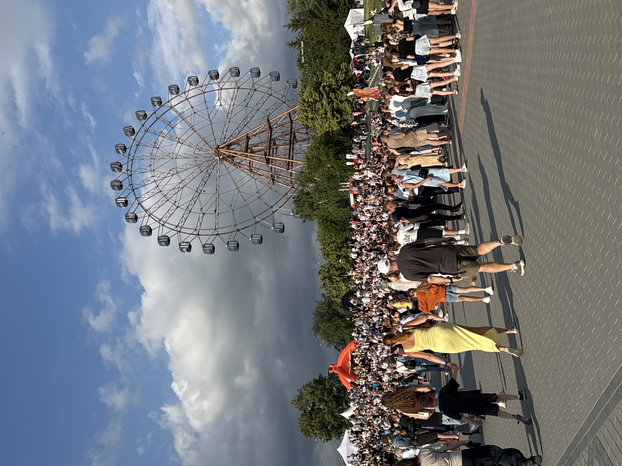 A large crowd of people are gathered in an outdoor space with a Ferris wheel in the background. The Ferris wheel is tall and appears to be made of metal, with many gondolas. In the middle of the crowd, there is a large, bright orange inflatable structure. The ground is paved with gray interlocking bricks. The sky is blue with some white and gray clouds. Trees are visible in the background, behind the crowd and Ferris wheel. The image appears to be taken during the daytime, possibly in the late afternoon or early evening, given the shadows cast by the people. The overall scene suggests an outdoor event or festival. This location is identified as the Novosibirsk Expoсenter in Novosibirsk, Russia.