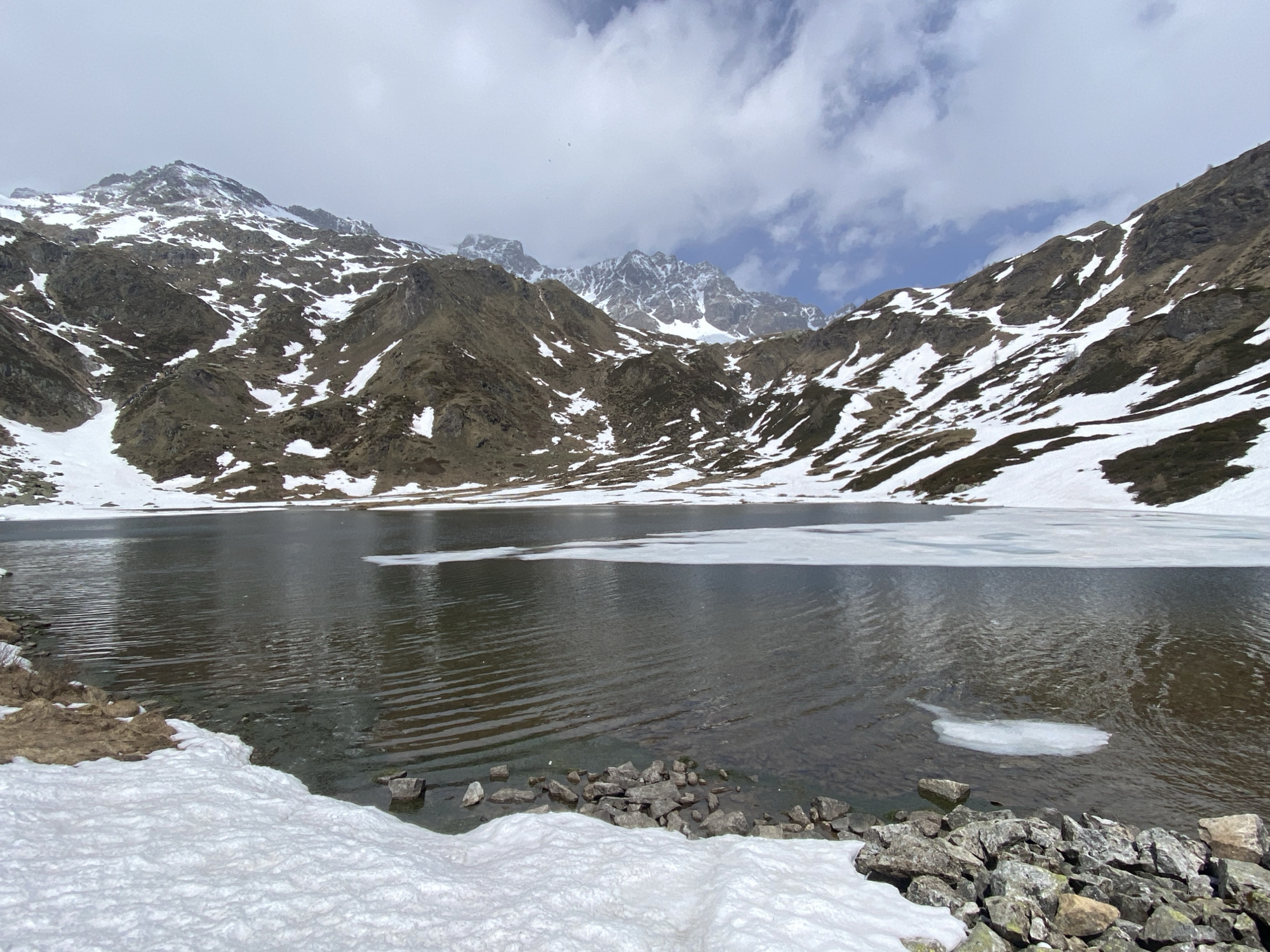This wide-angle photograph captures a majestic alpine landscape, centered on a large, dark lake nestled among rugged, snow-covered mountains. The lake's surface is dark and reflective, showing gentle ripples, and features several patches of floating ice, including a larger formation across the mid-distance and smaller fragments closer to the foreground.
The immediate foreground reveals a rocky shoreline partially covered in fresh snow, with some patches of dry, brown grass or moss on the left. Numerous grey and brown rocks line the water's edge on the right.
The surrounding mountains rise steeply from the lake, displaying a striking contrast between their dark, earthy rock and extensive patches of white snow. Snow streaks down many of the slopes, and the distant peaks are heavily snow-capped. The sky above is a mix of white and grey clouds, with occasional glimpses of pale blue, suggesting a partly cloudy day.
The scene depicts a natural, untouched environment, likely experiencing an early spring thaw, given the combination of persistent snow and visible bare ground. There are no people, structures, or any discernible activity. The overall impression is one of serene, cold, and powerful natural beauty. No text is visible in the image. The location is contextualized as Baceno, Italy.