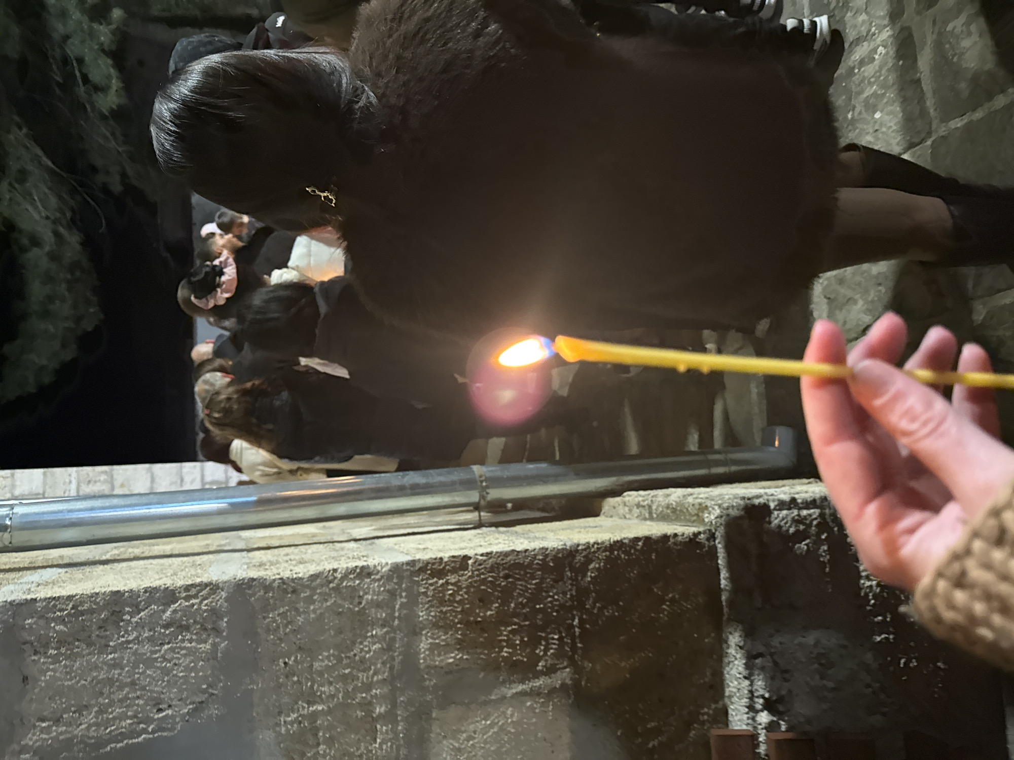 A slightly blurry, rotated image shows a person's hand holding a lit yellow taper candle, close to a low concrete wall capped with a silver metal pipe. The person wears a brown knit sweater. Behind the candle and wall is a group of people, including a woman with dark hair pulled back and wearing a dark, fur-like coat, standing near what appears to be a stone or concrete structure or fountain. Some greenery is visible on the left side of the image. The location is unknown and cannot be confirmed to be Byala Slatina, Bulgaria.