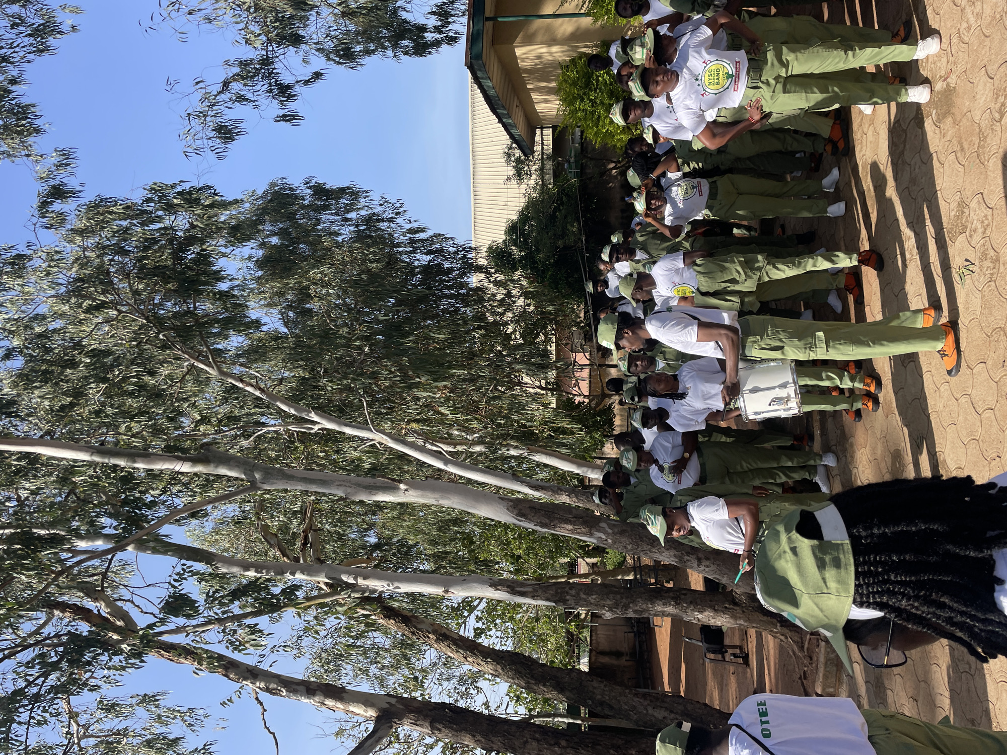 A group of young adults, dressed in matching green and white uniforms, are gathered outdoors in what appears to be a public space in Jos, Nigeria. The individuals are wearing khaki trousers and shirts, with some also wearing white t-shirts underneath, and green caps. Many are wearing the caps backwards or forwards, and some have their arms behind their backs. The group is composed of both men and women, and their faces show a range of expressions, from serious to smiling.
In the foreground, a young woman is holding a drum, suggesting a band or musical performance. Her stance indicates she is ready to play or is about to begin. Another individual next to her is also holding a drum. The ground is covered in red-brown earth, with some scattered leaves and shadows cast by the trees.
The environment is characterized by a cluster of tall, slender trees with smooth, pale bark, and their branches are covered in green leaves. In the background, there is a low-lying building with a light-colored, corrugated roof and yellow walls, suggesting it might be a school or administrative building. The sky is clear and blue, indicating a sunny day. The overall scene suggests an organized event or gathering, possibly a cultural or ceremonial occasion, taking place in a semi-arid environment typical of Jos.