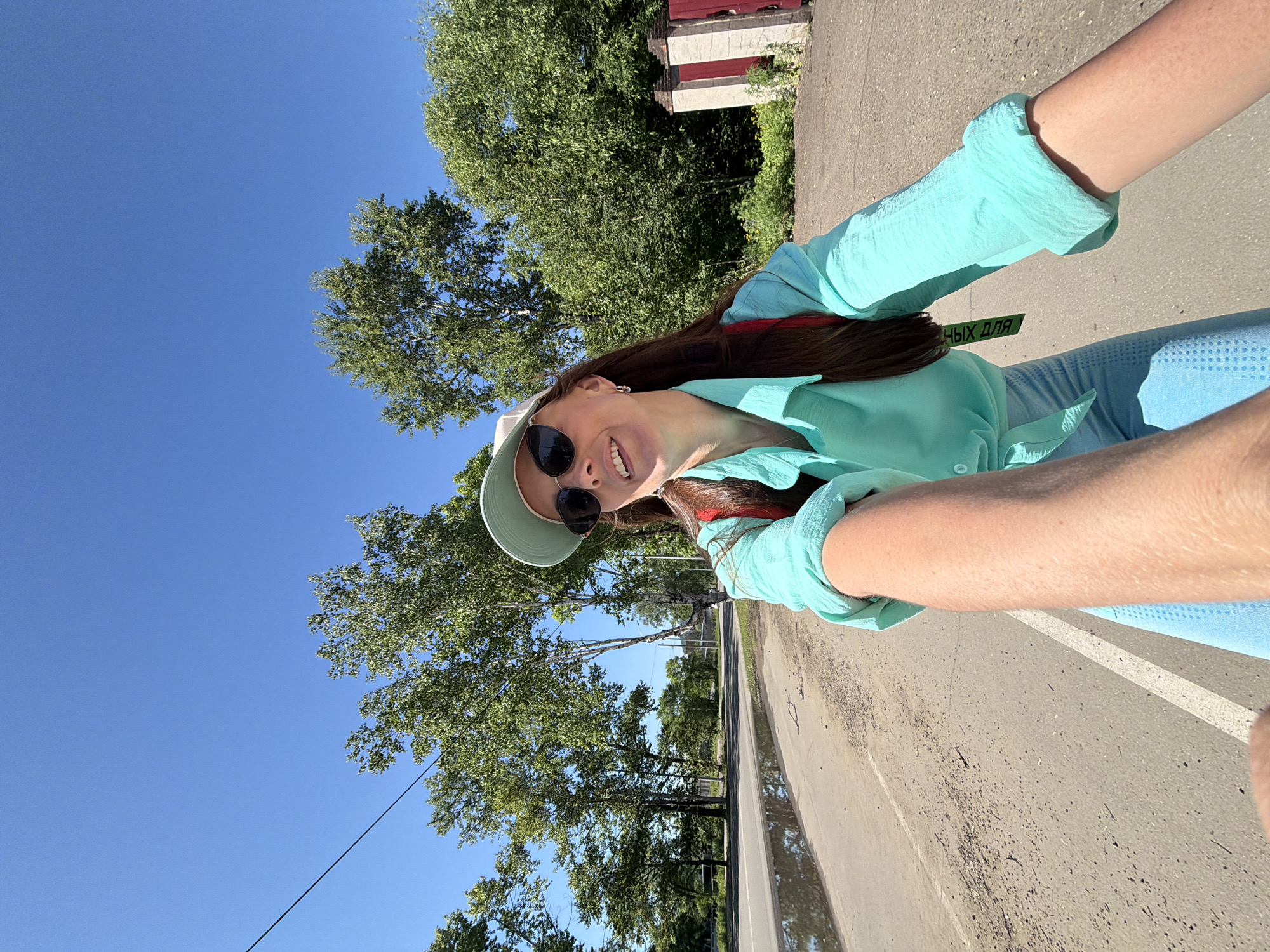 A full-body selfie taken from a low angle shows a woman with long brown hair wearing a light blue cap, sunglasses, and a turquoise shirt tied at the waist, with light blue leggings. She is smiling and looking upwards and to her right. A red backpack is visible on her shoulders. The background shows a tree-lined street with a paved road, a puddle of water on the left side, and a brick and white pillar fence on the right. The scene is set in Birobidzhan, Russia, under a clear blue sky.