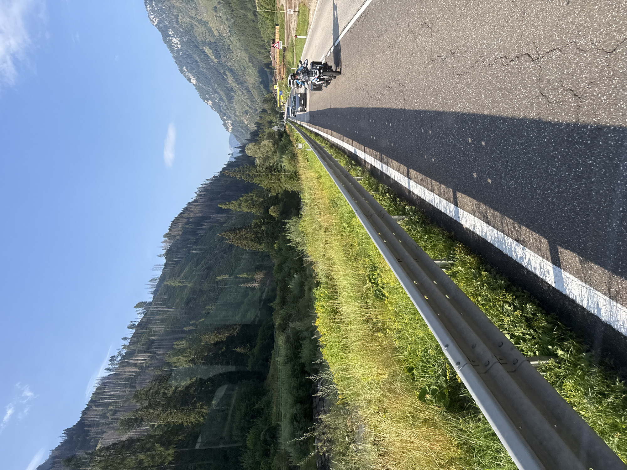 The image depicts a road winding through a valley, likely in the vicinity of Predazzo, Italy, given the mountainous terrain and lush vegetation. A motorcycle with a rider is prominently featured in the foreground, traveling on the right side of the asphalt road. The motorcycle is white and black with saddlebags. Several cars are visible further down the road, suggesting moderate traffic. The surrounding environment consists of steep, tree-covered mountainsides, with a clear blue sky overhead. The road is bordered by a metal guardrail and a grassy, vegetated area. The lighting indicates it is daytime, likely morning or afternoon.