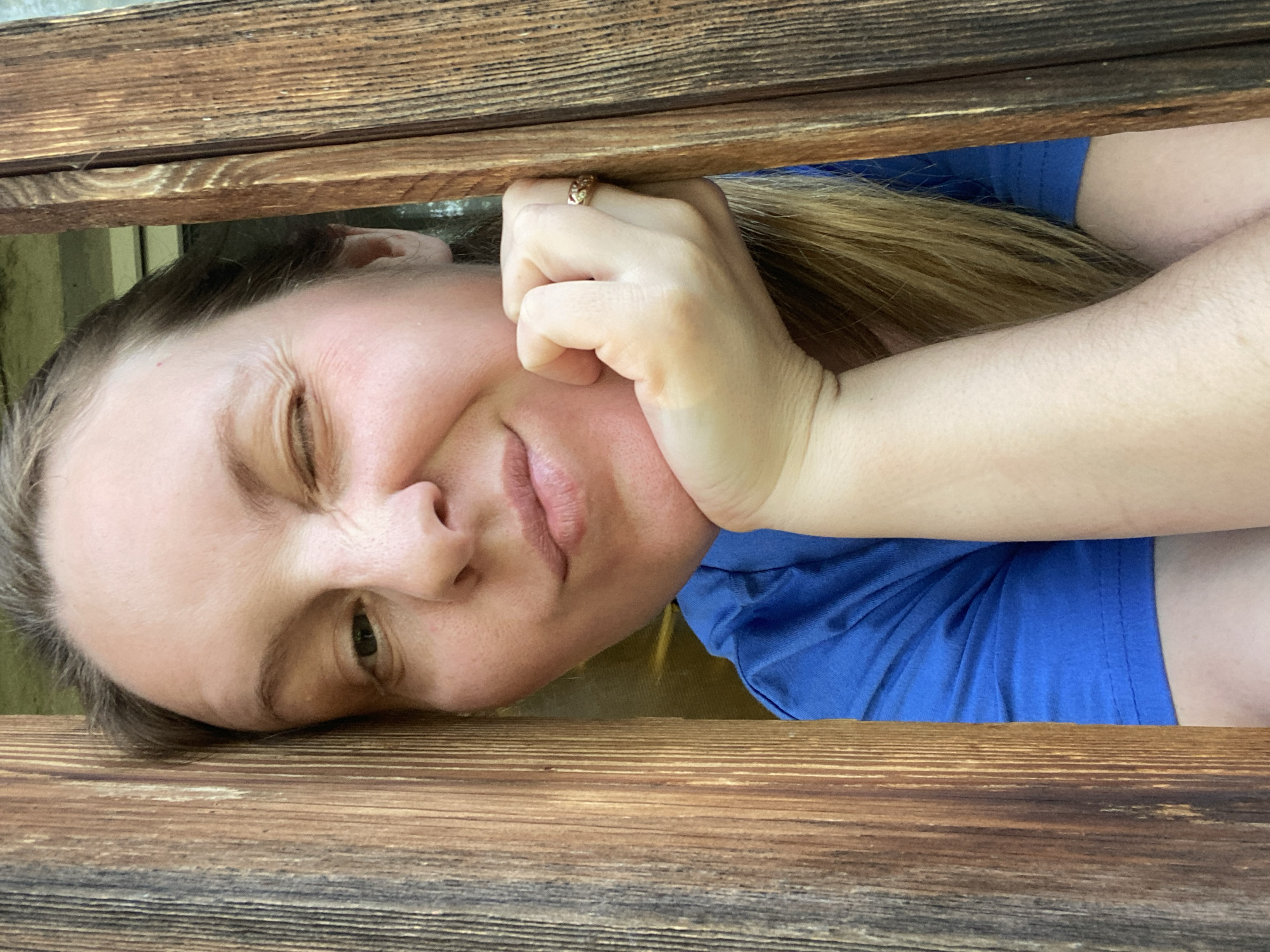 A woman wearing a blue t-shirt peers through a wooden structure, likely a fence or railing. Her face is close to the wood, with her left cheek resting against it. She is looking at the camera with one eye squinted and the other open, and her lips are pursed in a slight frown or pout. Her right hand is near her face, with a ring visible on one of her fingers. The background behind the woman is slightly blurry, suggesting a shallow depth of field. There is nothing to indicate this photo was taken in the city of Noviy, Russia.