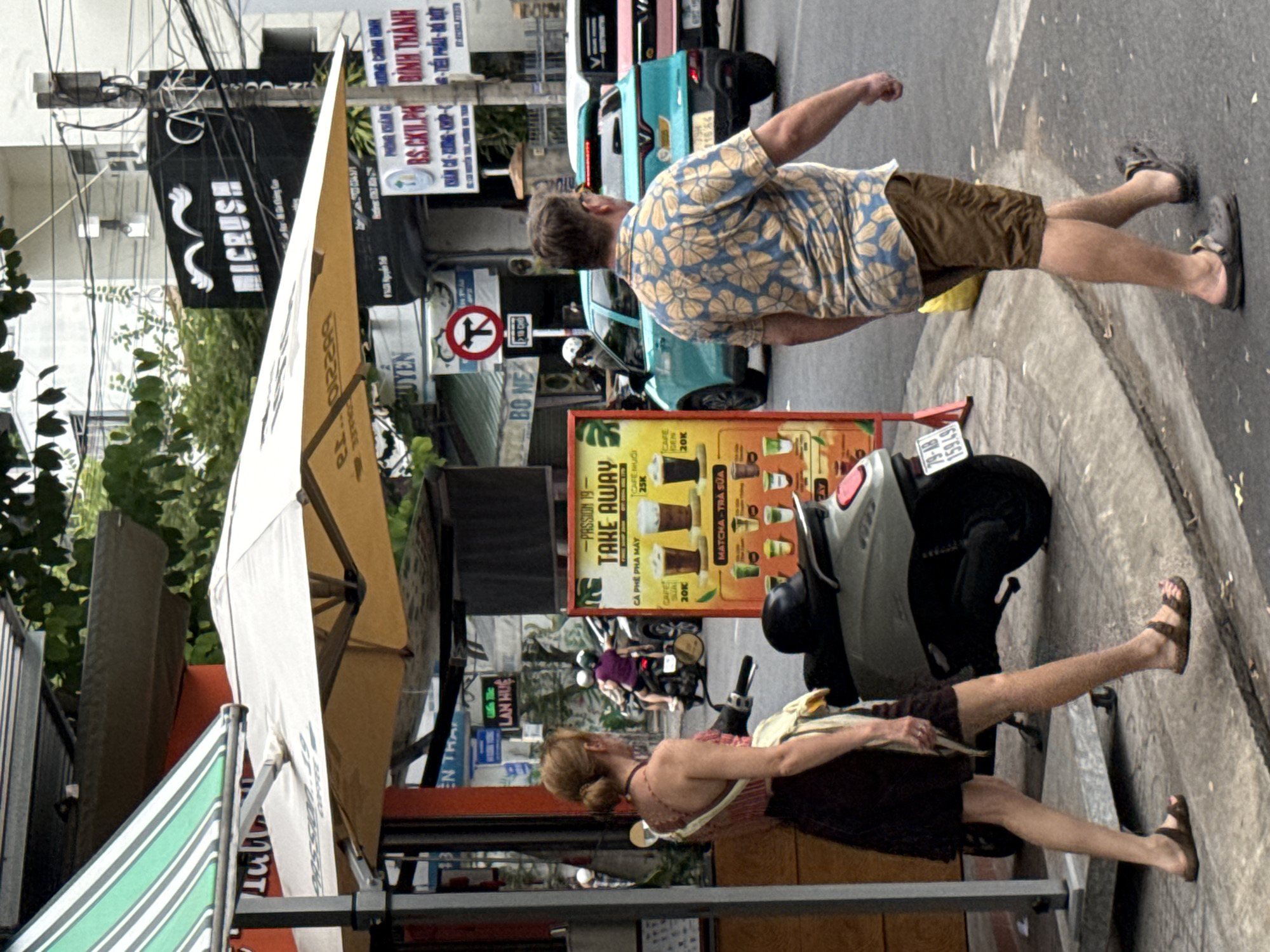The image depicts a street scene in Nha Trang, Vietnam, near the intersection of Tran Phu and Hung Vuong streets. In the foreground, a woman with blonde hair in a bun, wearing a patterned top and a dark skirt, walks away from the camera towards a scooter. To her right, a man with brown hair, wearing a blue and yellow floral shirt and brown shorts, walks in the same direction. Behind them, a teal SUV and a pink vehicle are visible on the street. A large yellow umbrella shades a table with a "TAKE AWAY" sign advertising coffee and matcha drinks. The background features buildings with signage, including a black sign with "MICRUSH" in white lettering, and overhead electrical wires. The pavement is wet, suggesting recent rain.