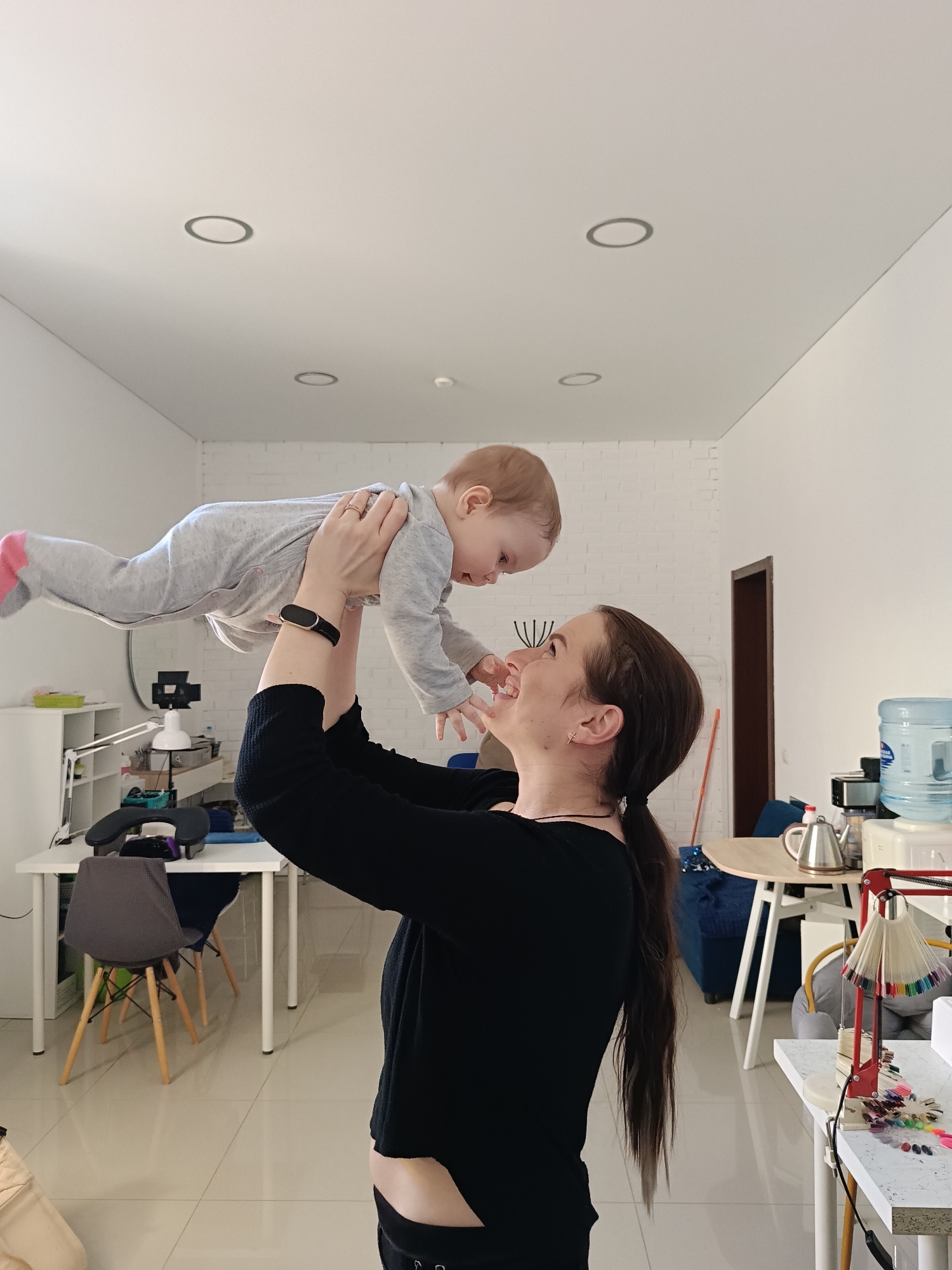 A woman is holding a baby up in the air inside a brightly lit room. The woman has brown hair pulled back in a ponytail and is wearing a black long-sleeved shirt. The baby is wearing a gray jumpsuit with pink socks. The room has white walls and a tiled floor. In the background, there are several tables and chairs, a water cooler, and a desk with a computer and other equipment. The scene is taking place in Chaikovsky, Russia.