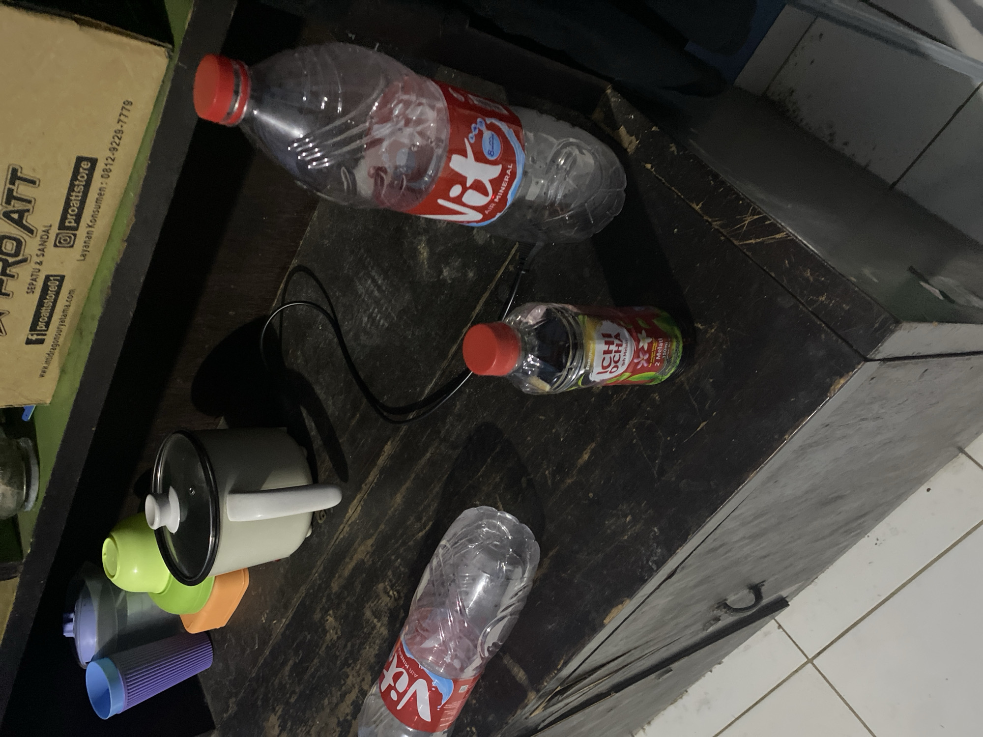 The image shows a cluttered wooden surface, possibly a shelf or table, in what appears to be a domestic setting in Tangerang, Indonesia. On the left, there are several items including a purple ribbed cup, a light blue lidded container, an orange rectangular object, and a green bowl. A small white pot with a glass lid and a white handle is also present. In the center, a small bottle of "ICHIOCHA Teh Melati" is visible. To the right, a large clear plastic bottle of "Vit Air Mineral" is positioned, with its red cap slightly askew. A black electrical cord is visible behind the bottles. The background is dark and out of focus, with a cardboard box containing text that includes "PRO ATT SEPATU & SANDAL" and a phone number for customer service. The wooden surface is worn and scratched, suggesting regular use. The lighting is dim, contributing to the overall dark atmosphere.