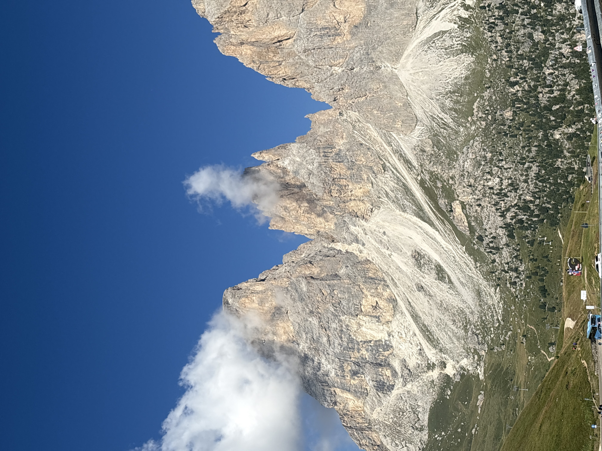 The image depicts the Sassolungo group in the Dolomites, a mountain range located in northern Italy, near the municipality of Canazei. The iconic peaks of Sassolungo and Sassopiatto dominate the upper half of the frame, their jagged, rocky faces bathed in sunlight under a clear blue sky. Wisps of white clouds cling to the upper reaches of the mountains, partially obscuring some of the sheer rock faces.
In the foreground, a verdant green slope descends, dotted with scattered dark green pine trees and rocky outcrops. Several winding paths are visible snaking up the mountainside, indicating hiking trails. A paved road runs horizontally across the lower part of the image, with a blue bus parked on it. Further along the road, a white car is parked. People are visible on the road and the lower slopes, appearing to be engaged in recreational activities. A large, circular billboard with cartoonish figures is also visible near the road. The overall impression is a scenic mountain landscape on a bright, sunny day.