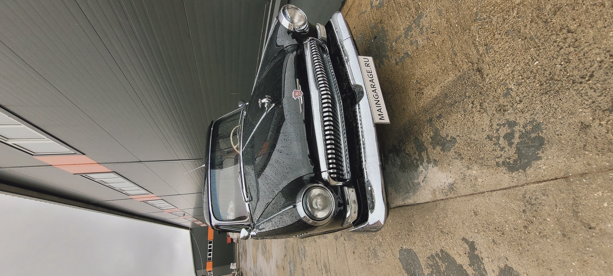 A vintage black car is parked in front of a building in Unknown, Unknown. The vehicle, seemingly a classic model, is positioned on a gray concrete surface. The building behind the car has a dark gray facade with a pattern of horizontal lines. The sky is overcast, and there are rain droplets visible on the car's surface. A sign with "MAINGARAGE.RU" is mounted on the front of the car.