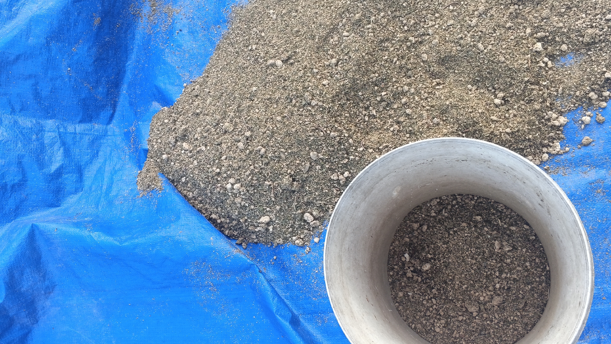 A pile of granular material, possibly soil or compost, is spread across a bright blue tarp. To the right of the pile, a silver cylindrical container, appearing to be made of metal, is positioned on the tarp. The container is partially filled with the same granular material as the pile. The scene is observed from a top-down perspective. There are no people or animals visible in the image. The location is identified as Soko, Indonesia.