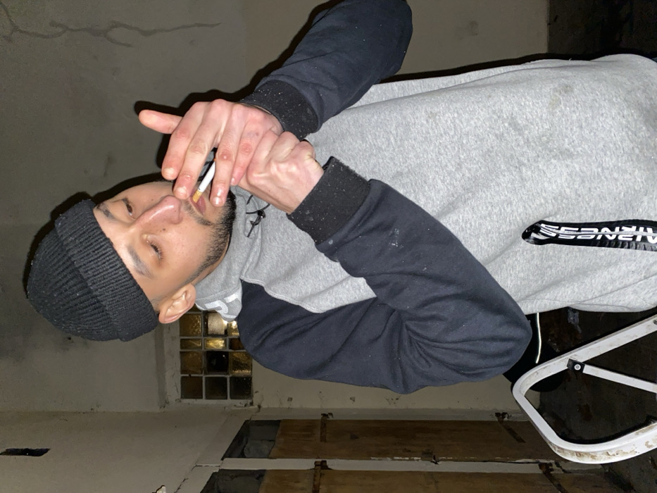 A man wearing a black beanie and gray and black hoodie is smoking a cigarette. He has a beard and mustache. He is standing in a room with a broken floor and a window with textured glass. A white ladder is visible in the background. The lighting is dim.