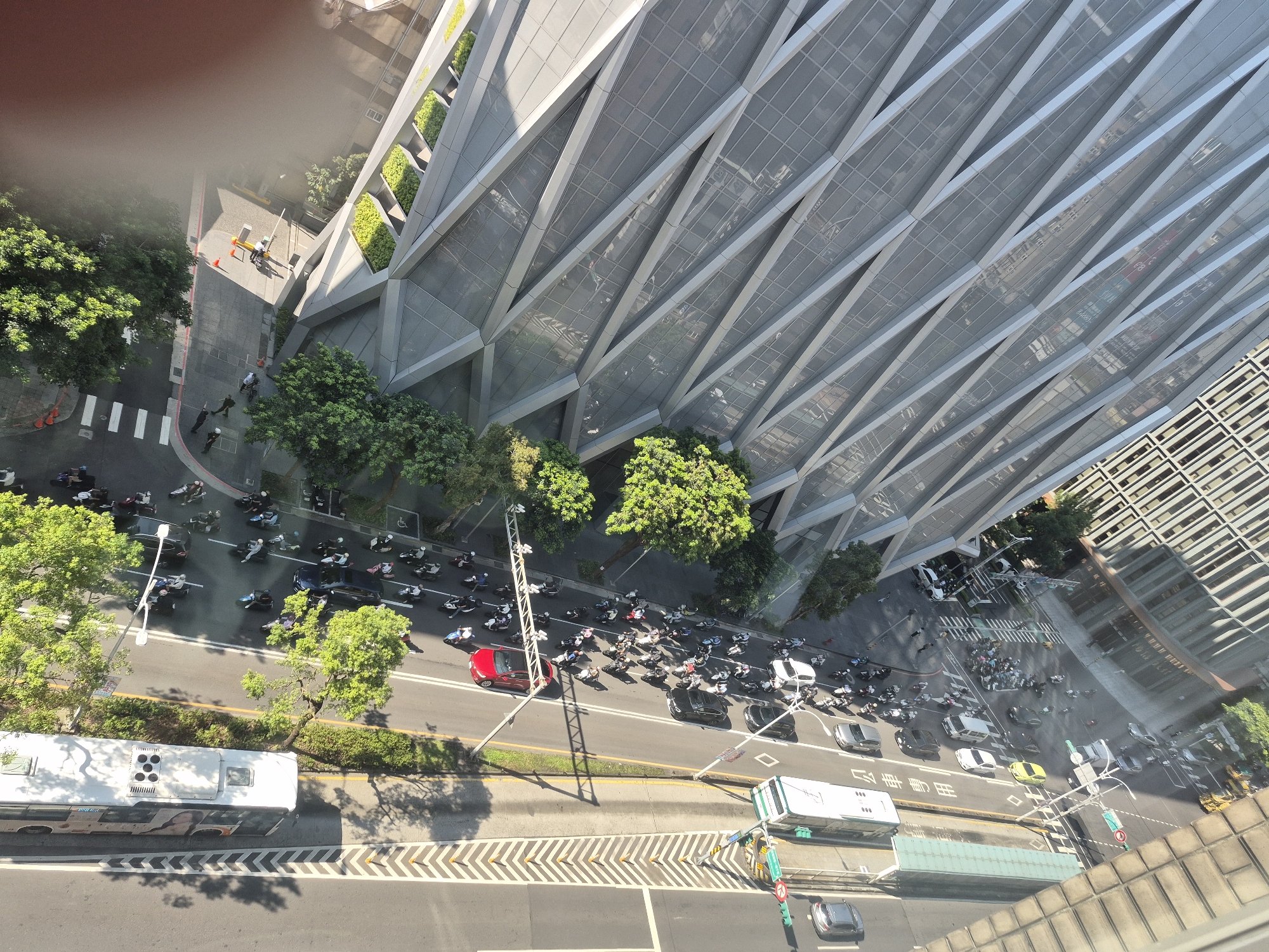 An aerial, high-angle shot looks down on a busy intersection and street in Taipei, Taiwan. The foreground features the top of a white bus with several windows and a light colored roof, parked on the left side of the frame. The bus has a black and white chevron pattern on the road in front of it. To the right of the bus is a street with two lanes of traffic moving away from the viewer. The lanes are separated by a yellow line. On the right side of the street, a sidewalk and green foliage line the road.
Further up the street, a dense cluster of motorbikes and scooters are stopped at what appears to be a traffic light or intersection. Many of the riders are wearing helmets. Several cars are also present, including a red sedan, a black SUV, and a white car. A tall, modern building with a distinctive angled glass facade dominates the right side of the frame, casting shadows onto the street below. The building has a repeating geometric pattern of triangular shapes and recessed green planters. Across the street from this building, another, older building with a grid-like facade is visible. The overall scene depicts urban traffic and architecture in Taipei under bright daylight.