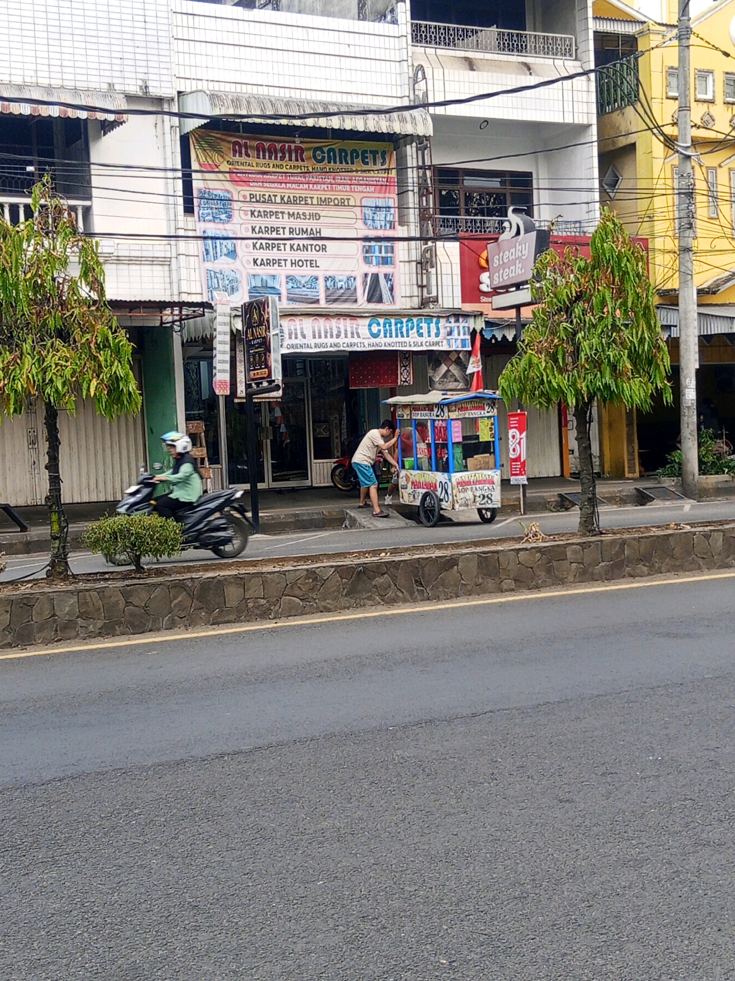 The image depicts a street scene in Lubuklinggau, Indonesia. A person is riding a dark-colored motorcycle from left to right, wearing a green shirt and a helmet. In the background, a food cart is visible with two individuals nearby. The cart is decorated with colorful signage and displays prices, including "28" and "81". To the right of the food cart, a "Steaky Steak" sign is prominent. The buildings behind the street feature various shop fronts, including one with a banner for "AL NASIR CARPETS". The environment includes a paved road, a stone wall bordering the sidewalk, and mature trees with green foliage. The buildings appear to be multi-story structures with a mix of modern and older architectural elements. The lighting suggests it is daytime.