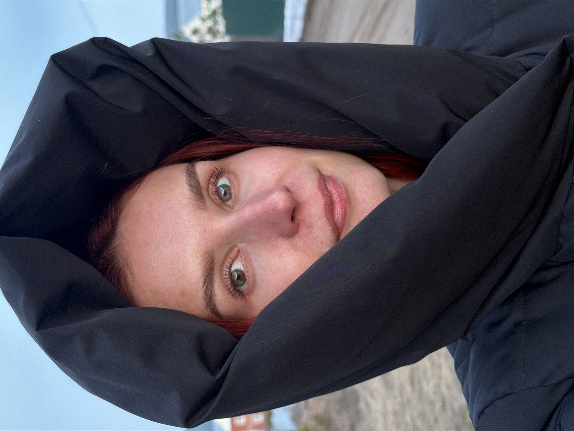 A close-up, tilted shot of a woman with fair skin, red hair, and light green eyes, with a slight smile. She is wrapped in a large, black hooded jacket that almost completely covers her head. The background suggests an outdoor location, possibly in the city of Chaikovsky, Russia, with a glimpse of the sky, a beach, and greenery in the distance.