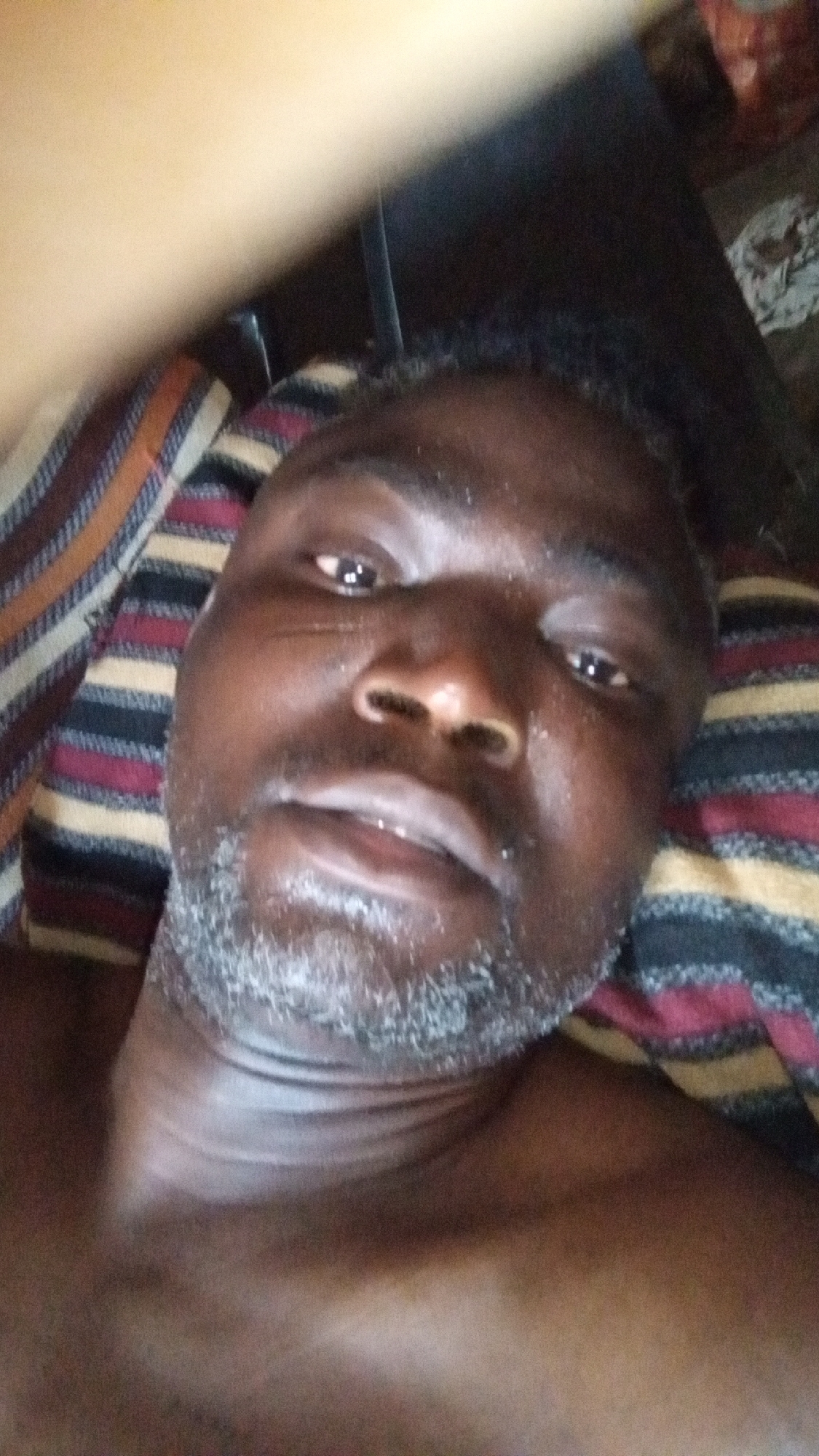 The image is a close-up, low-angle portrait of an adult individual, likely male, with dark skin, lying down and looking directly at the camera. The person has short, dark hair and a salt-and-pepper beard or stubble. Their facial expression is neutral.
The individual's head is resting on a striped pillow or blanket, visible along the left and right sides of the frame. The fabric features horizontal stripes in shades of dark red, black, and light brown or tan. A blurred, overexposed, light-colored object or body part extends from the top left corner across the upper portion of the image, partially obscuring the background. The background is otherwise dark and indistinct, suggesting an indoor setting. The person's skin appears somewhat glossy, possibly from natural oils or perspiration. No specific activities, time of day, weather conditions, or textual elements are visible.