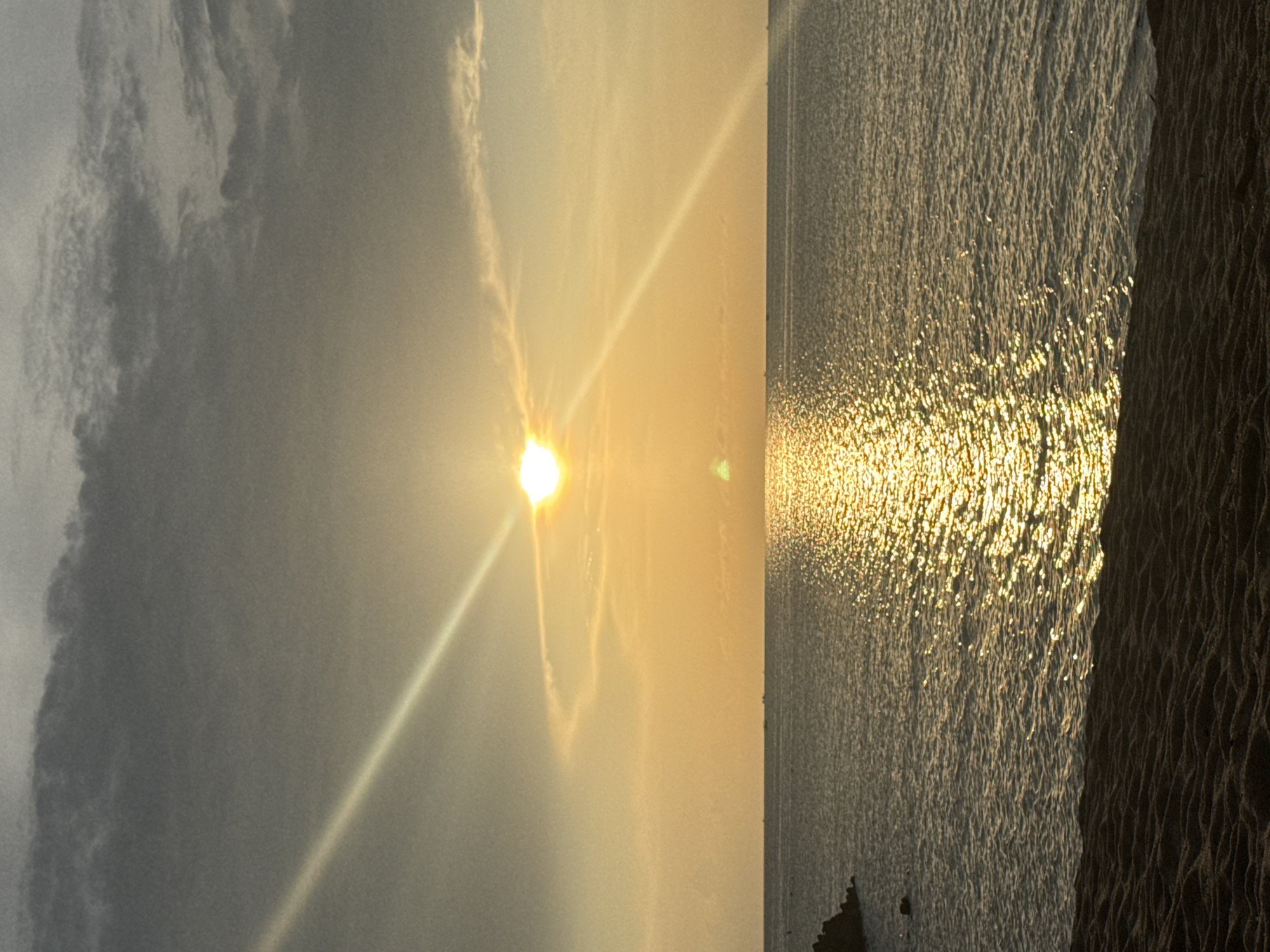 The image depicts a sunset view in Baan Wang Ta Kien, Thailand. The sun is low in the sky, casting a golden glare across the water. The sky is partially covered with clouds. The water surface is rippled, reflecting the sunlight. A shoreline or embankment is visible on the right side of the image.
