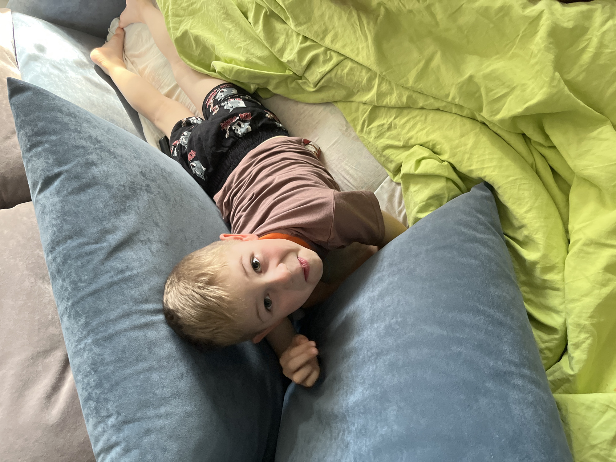 This image captures an indoor domestic scene in Rusanovo, Russia. A fair-skinned young child with short blonde hair lies prone on a light-colored sheet. The child wears a brownish-pink short-sleeved t-shirt and black shorts featuring a repeating pattern, possibly depicting cartoon characters. The child's gaze is directed towards the camera with a slight, mischievous expression. Their right arm is bent, with the hand resting on a large, plush blue velvet pillow that occupies the foreground. Another similar blue pillow is positioned behind the child's head, bordering a light brown upholstered surface in the upper background. A bright lime green duvet or blanket is draped and crumpled across the right side of the frame, partially covering the light sheet. The scene is brightly lit, highlighting the textures of the various fabrics.