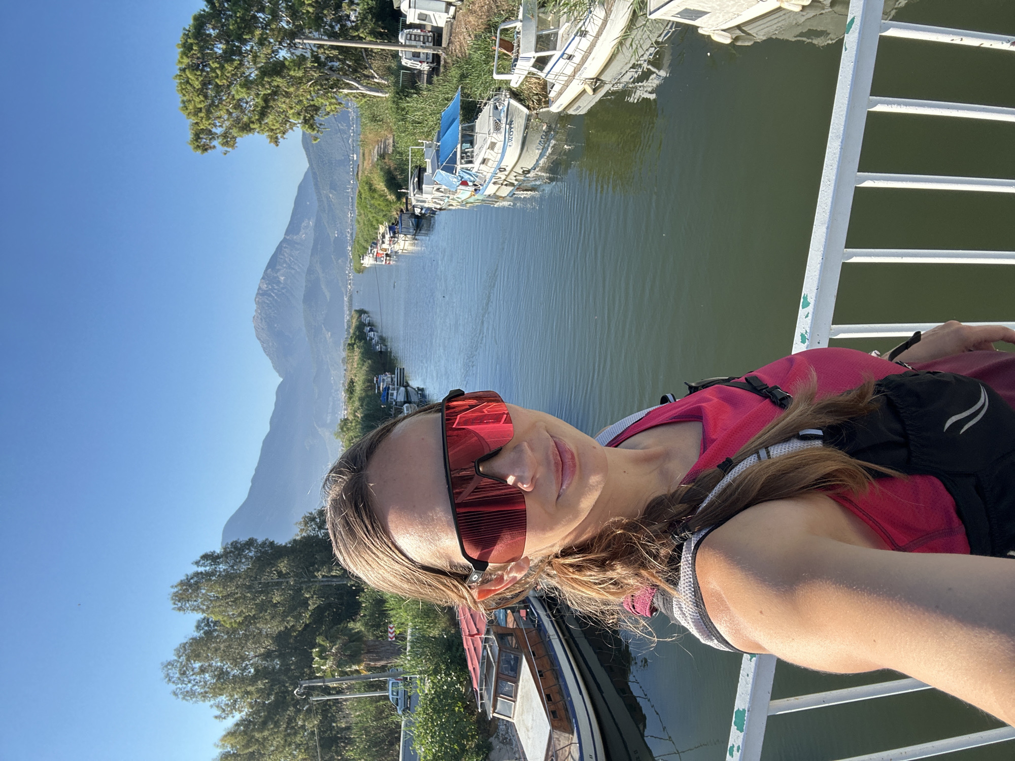 The image is a selfie of a woman standing on a bridge. She is wearing sunglasses, a pink tank top, and a backpack. The background shows a view of a river with boats docked on either side, a mountain in the distance, and trees. The city of Fethiye, Turkey is visible. The bridge has white metal railings. The water in the river is a dark green color. The sky is clear and blue.