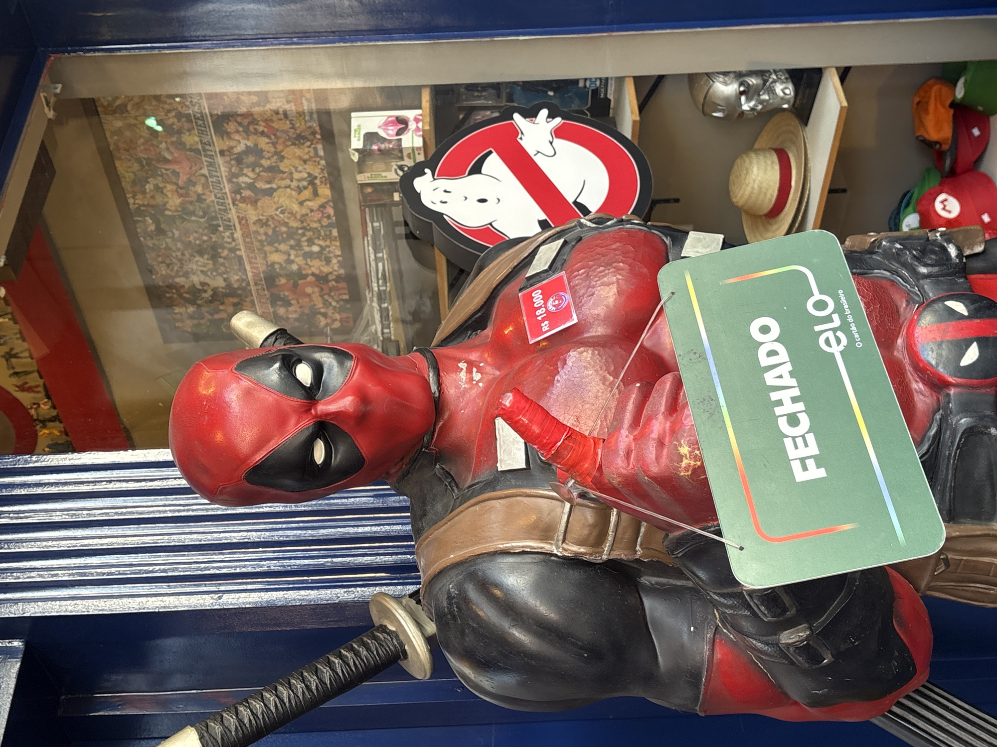 A life-sized Deadpool statue stands in front of a shop window in São Paulo, Brazil. The statue is dressed in its iconic red and black suit and holds a green sign that reads "FECHADO" (Closed) in white letters. The sign also features the "ELO" logo, indicating a Brazilian payment method. Behind the statue, the shop window displays various items, including framed posters, action figures, hats, and other merchandise. A "Ghostbusters" logo is also visible in the window. The shop appears to be an establishment selling collectibles or novelty items. The background consists of a blue, ribbed surface on the left and the shop interior on the right.
