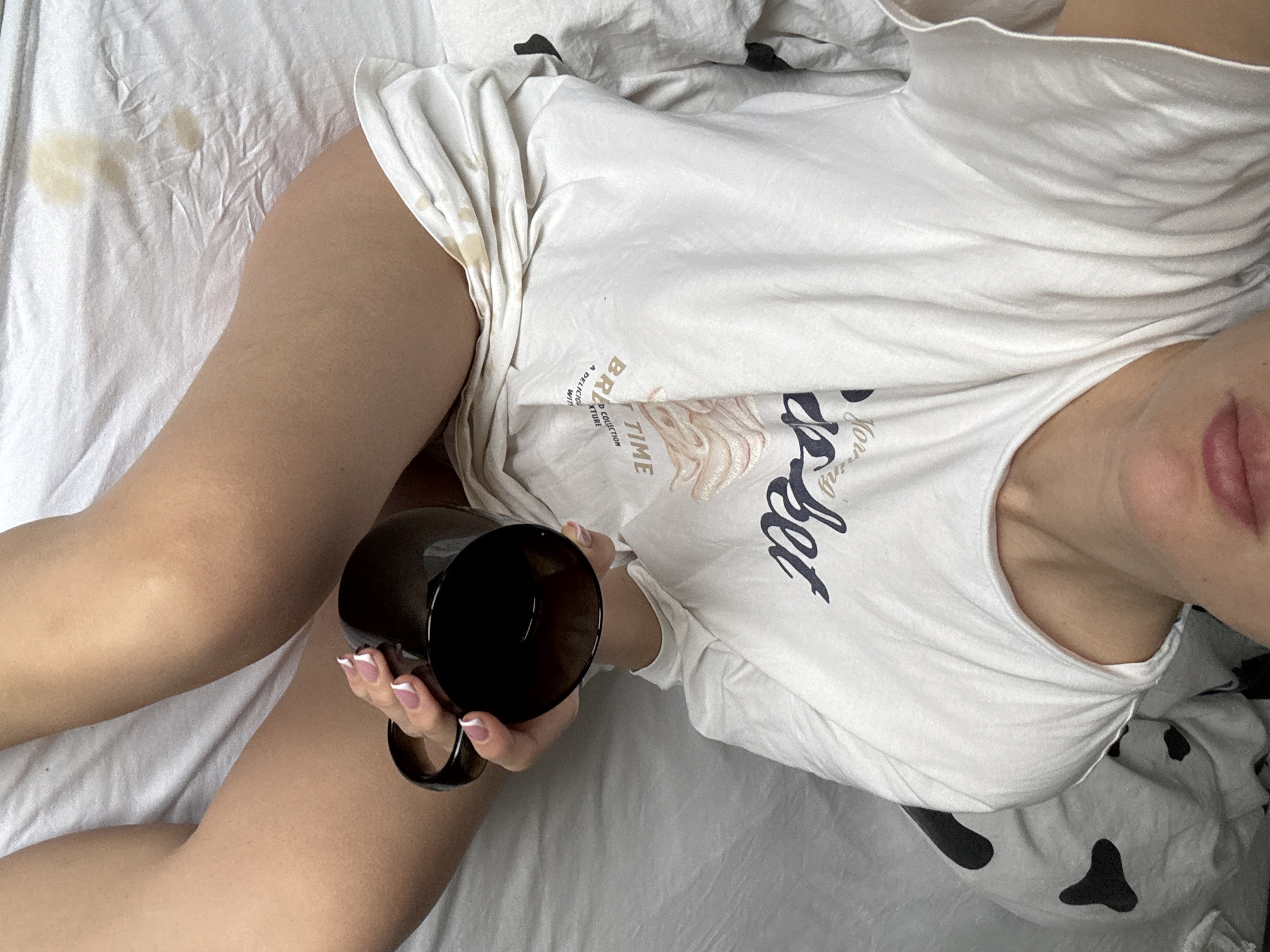 The image presents a top-down view of a person relaxing in bed, holding a dark mug. The individual is largely obscured by a white t-shirt and white bedsheets, with their legs and one arm visible. The scene suggests an indoor setting, likely a bedroom, without specific geographical indicators, though the location context is Chaikovsky, Russia.
The person is wearing a loose-fitting white t-shirt that appears to have graphics and text. Partially visible text on the shirt includes "BR" and "TIME," along with fragments like "A DELICIOU," "D COLLECTION," and "TEXTURE." Another section of the shirt displays stylized script that seems to say "Morning" or a similar word. The person's right hand, with manicured nails featuring white tips, holds a black or very dark brown mug.
The legs are uncovered, resting on the white bedsheets. There are noticeable light brown stains on the white bedsheet, particularly in the upper left corner, and similar subtle discoloration is visible on the t-shirt, suggesting a spill of a beverage like coffee or tea. A patterned pillow or duvet cover, featuring black, abstract shapes on a white background (resembling a cow print), is visible beneath the person's head/upper body on the right side of the frame. Only the lower part of the person's face, specifically the mouth with slightly parted, light-colored lips, is visible on the right side, offering no clear emotional cues. The overall ambiance suggests a casual, relaxed moment, possibly in the morning.