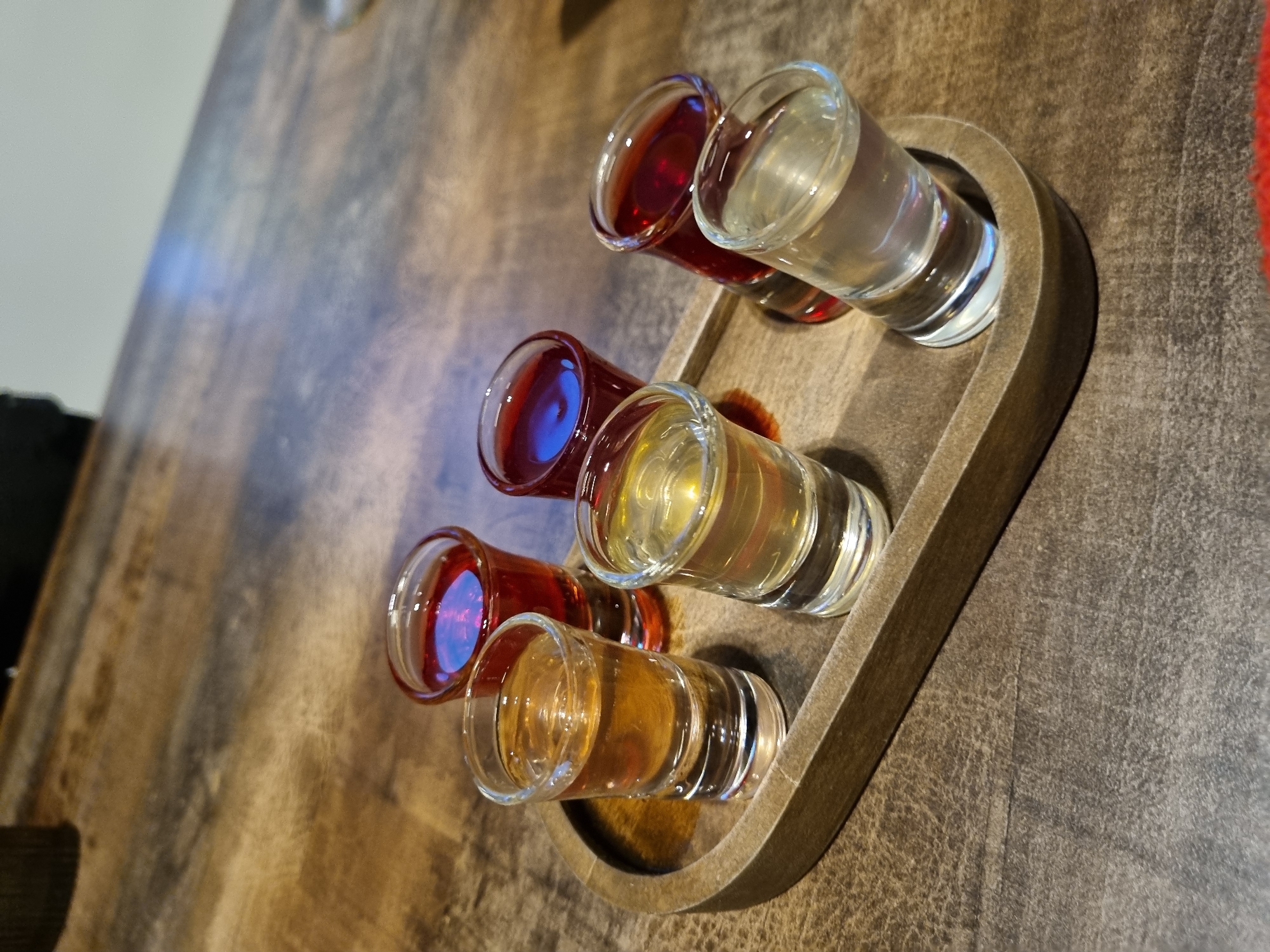 The image, taken in Chaikovsky, Russia, showcases six shot glasses meticulously arranged on a dark brown wooden serving tray. The tray, characterized by its smooth, slightly elevated edges and rounded corners, is positioned on a dark, textured wooden table with visible grain patterns. The ambient lighting causes bright reflections on the glass surfaces and the liquids within.
Each shot glass contains a distinct liquid, varying in color and hue. Starting from the top right and moving left across the rows: the first glass holds a clear, colorless liquid. Adjacent to it, the second glass contains a rich, dark red liquid. In the middle row, the glass on the right is filled with a golden-yellow beverage, while its counterpart on the left holds a vibrant reddish-purple liquid. The bottom row features a glass with an amber-brown liquid on the right, and the final glass on the left contains a bright red liquid, showing blue reflections from the surroundings.
The presence of multiple, varied beverages in shot glasses strongly suggests a tasting event or sampling session. The scene appears to be set indoors, possibly in a bar, restaurant, or a private residence, indicated by the table and the controlled lighting. There are no individuals visible in the frame, nor is there any discernible text. The composition focuses solely on the colorful display of drinks.