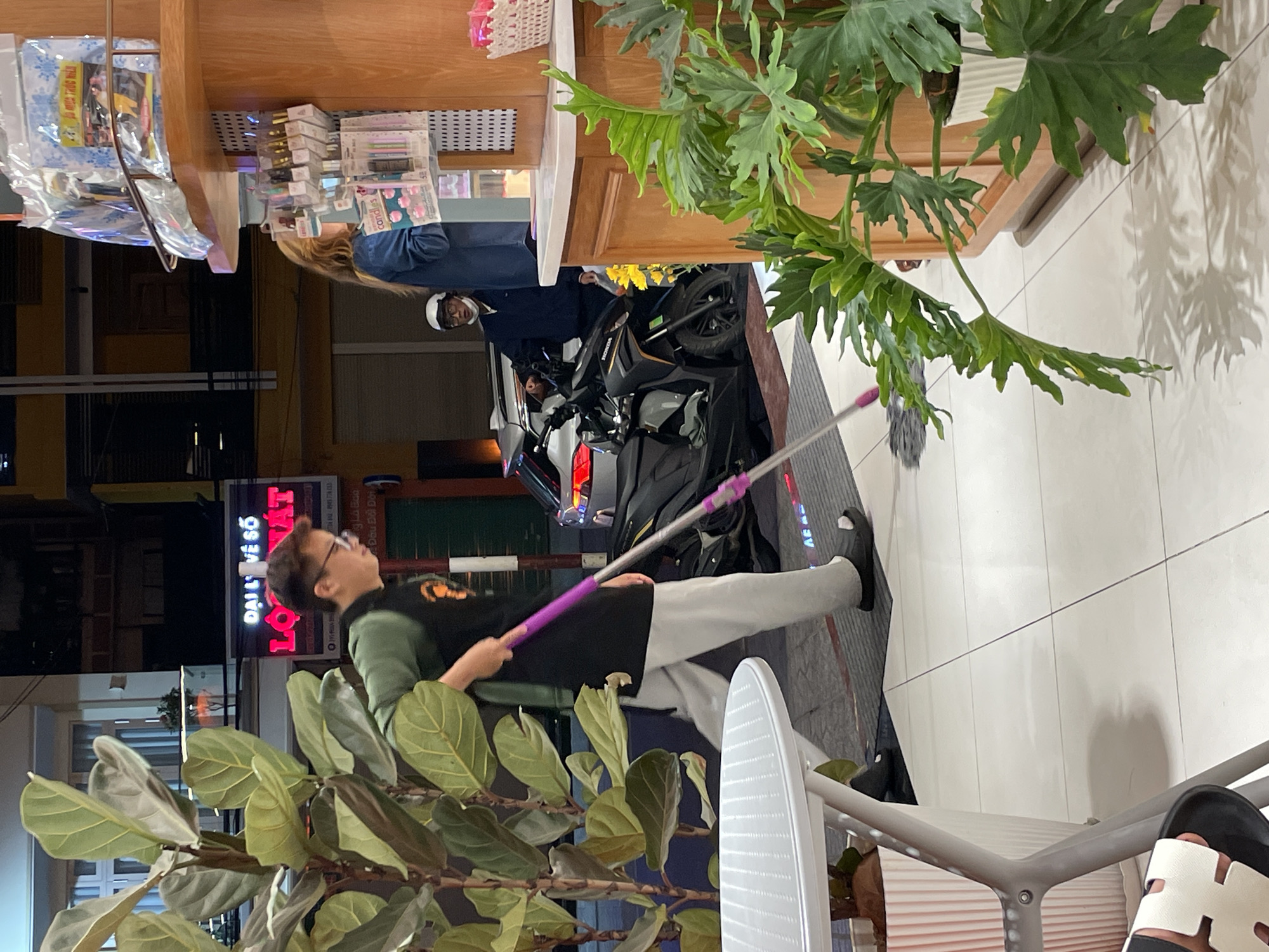 A young man, wearing glasses and a dark vest over a green shirt and light-colored pants, is mopping the tiled floor with a purple-handled mop outside a shop. A large potted plant with broad leaves is to his left. In the background, a woman in a blue jacket stands behind a display of goods, and a man wearing a white helmet and a dark jacket is near a motorcycle. A neon sign for "LO XAT" is visible further in the background. The scene appears to be on a street in Da Lat, Vietnam, likely in the evening, judging by the lighting and the dark sky.