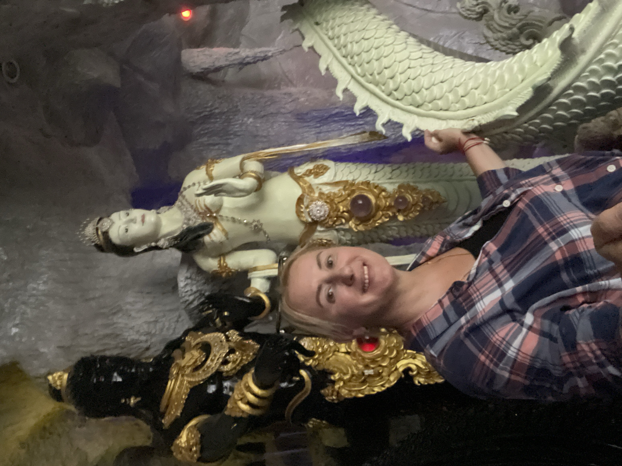 A woman with blonde hair is smiling as she poses for a photograph in front of two statues and a dragon sculpture. The woman is wearing a blue, pink, and white plaid shirt over a black top. To her left is a tall, black statue with gold ornate details, wearing a gold crown. To her right, and slightly behind, is a pale statue of a woman with a crown and elaborate jewelry. To the woman's right and in the foreground is a pale green sculpture of a dragon's body. The scene appears to be inside a cave or grotto, with rocky walls and artificial lighting, possibly in a temple or tourist attraction in Bo Win Subdistrict, Thailand.