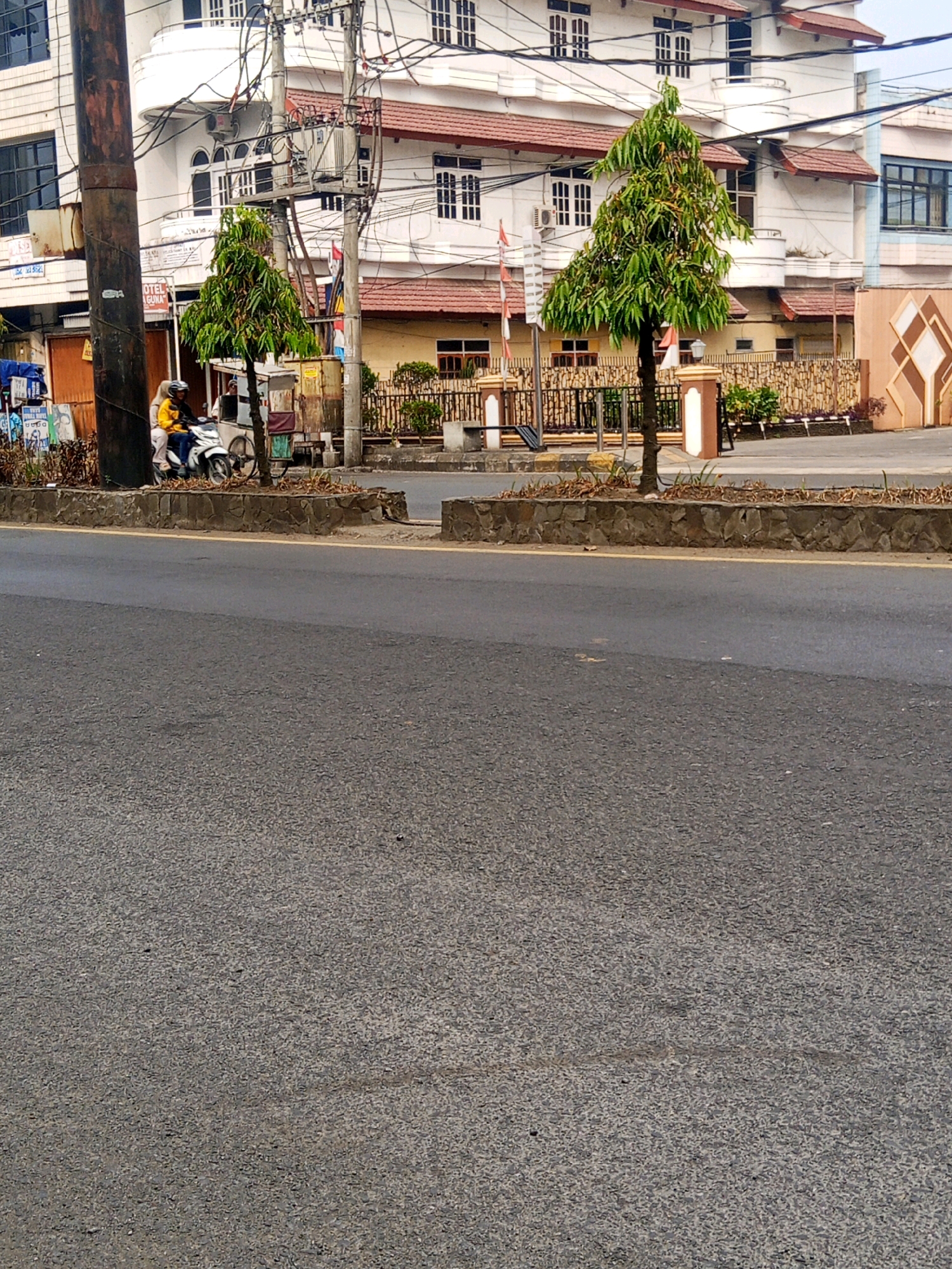 A street scene in Lubuklinggau, Indonesia, features a two-story building with a white facade, red tiled roofs, and numerous windows. In front of the building, a low stone wall separates a sidewalk from the street. Two conical trees are planted on the sidewalk, and Indonesian flags are visible behind them. A person on a motorcycle, wearing a yellow jacket and a helmet, is stopped near the left side of the frame, next to a cart with wheels. A utility pole with wires stands on the left, and a concrete structure with a checkered pattern is visible on the far right. The street in the foreground is paved with asphalt and has a yellow line marking the edge.