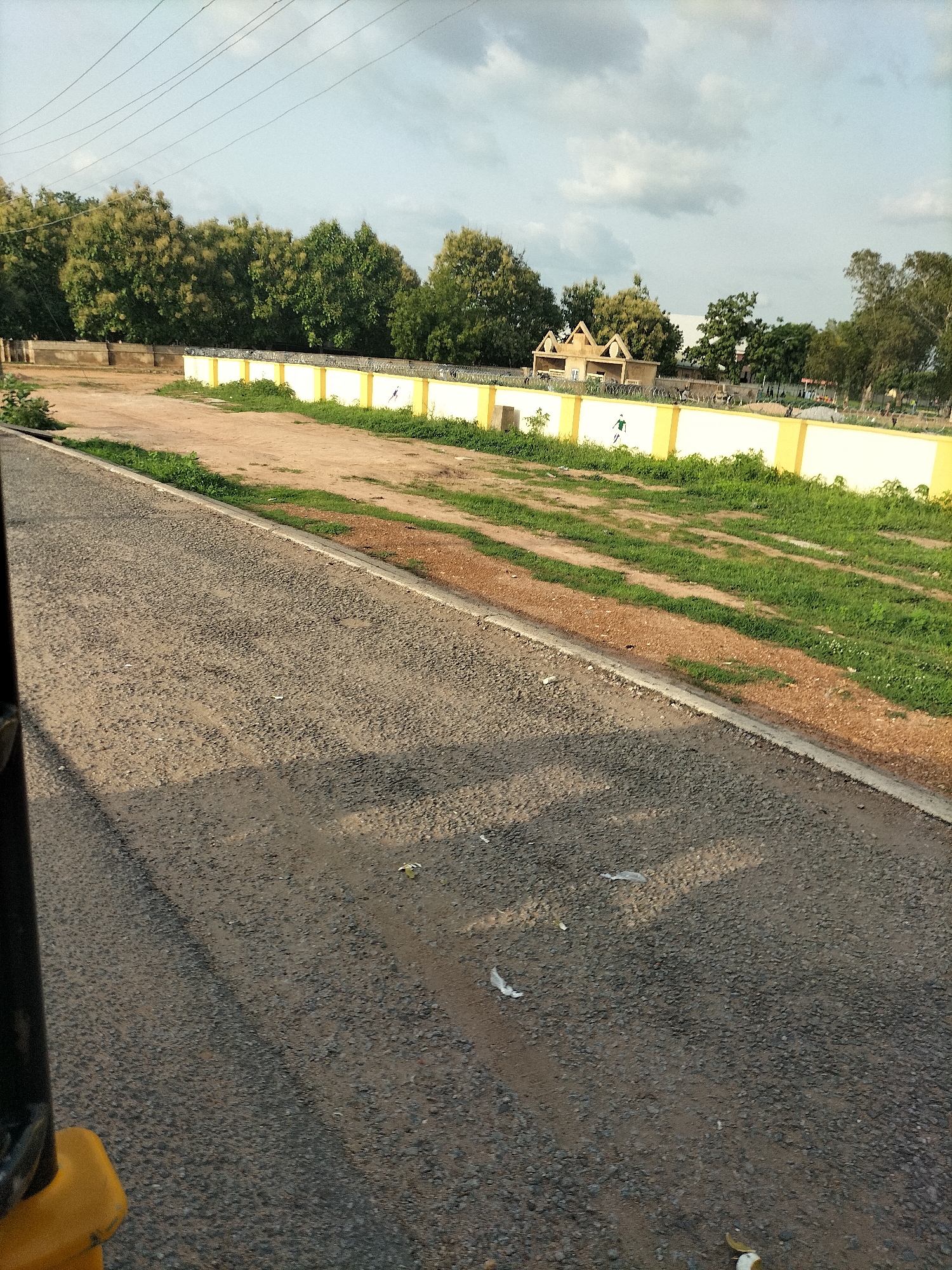 The image depicts an outdoor scene in Jalingo, Nigeria, focusing on a low, yellow-and-white wall bordering a field. The wall has a decorative pattern of what appear to be small spikes or ornamental finials along its top. On the wall, a stylized green figure resembling a soccer player is painted. Beyond the wall, several trees are visible, with some exhibiting a feathery or wispy foliage. In the background, a small, light-colored structure with a peaked roof is present, suggesting a park or recreational area. The ground in front of the wall consists of dirt and patches of grass. In the foreground, a paved road or pathway is visible, with tire tracks and scattered debris, including pieces of white paper. The sky above is partly cloudy. The overall impression is of a public space undergoing development or maintenance, with elements of sports and natural landscaping.
