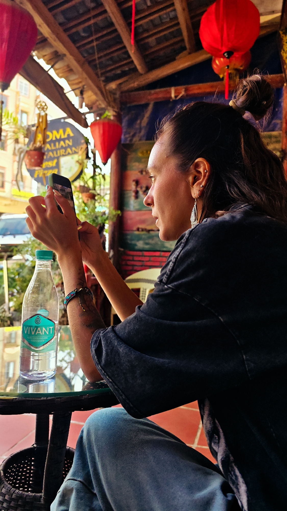 A woman is seated at a table in an open-air restaurant, holding a smartphone in her hands. She has dark hair pulled back into a bun, wears dangling earrings, a dark t-shirt, and blue jeans. A bottle of water labeled "Vivant" sits on the table in front of her. The restaurant appears to be located in Đồng Văn Commune, Vietnam, given the signs in the background and the style of architecture. The restaurant has red lanterns hanging from the ceiling and colorful decorations on the walls. Outside the restaurant, there is a street scene with buildings and vehicles.