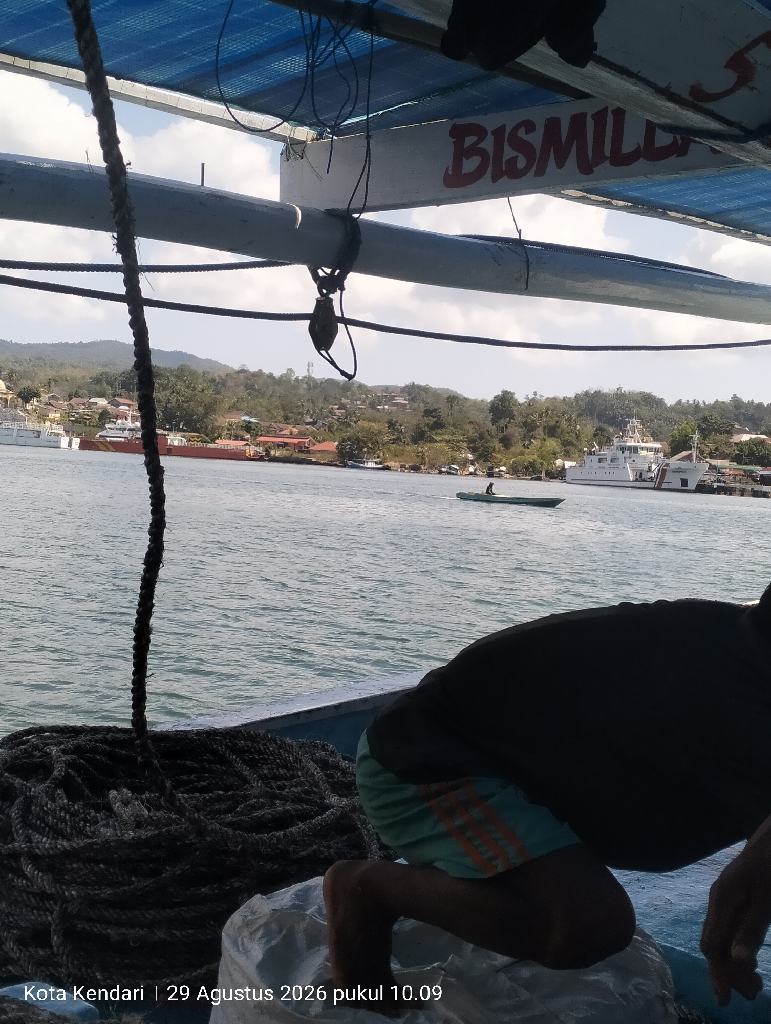 The image shows a view from a boat in Kendari, Indonesia, on August 29, 2026, at 10:09 AM. The foreground features a coil of thick rope on the deck of a boat, with a person crouching in the lower right corner, partially obscured by shadow. A large white structure, possibly a sail or canopy, is visible overhead. In the middle ground, the ocean is calm, with a small, narrow boat carrying a single person moving across the water. Further in the background, a larger white vessel, likely a ship, is docked near a coastline lined with buildings and lush green hills. The overall scene depicts a coastal environment with active maritime activity.