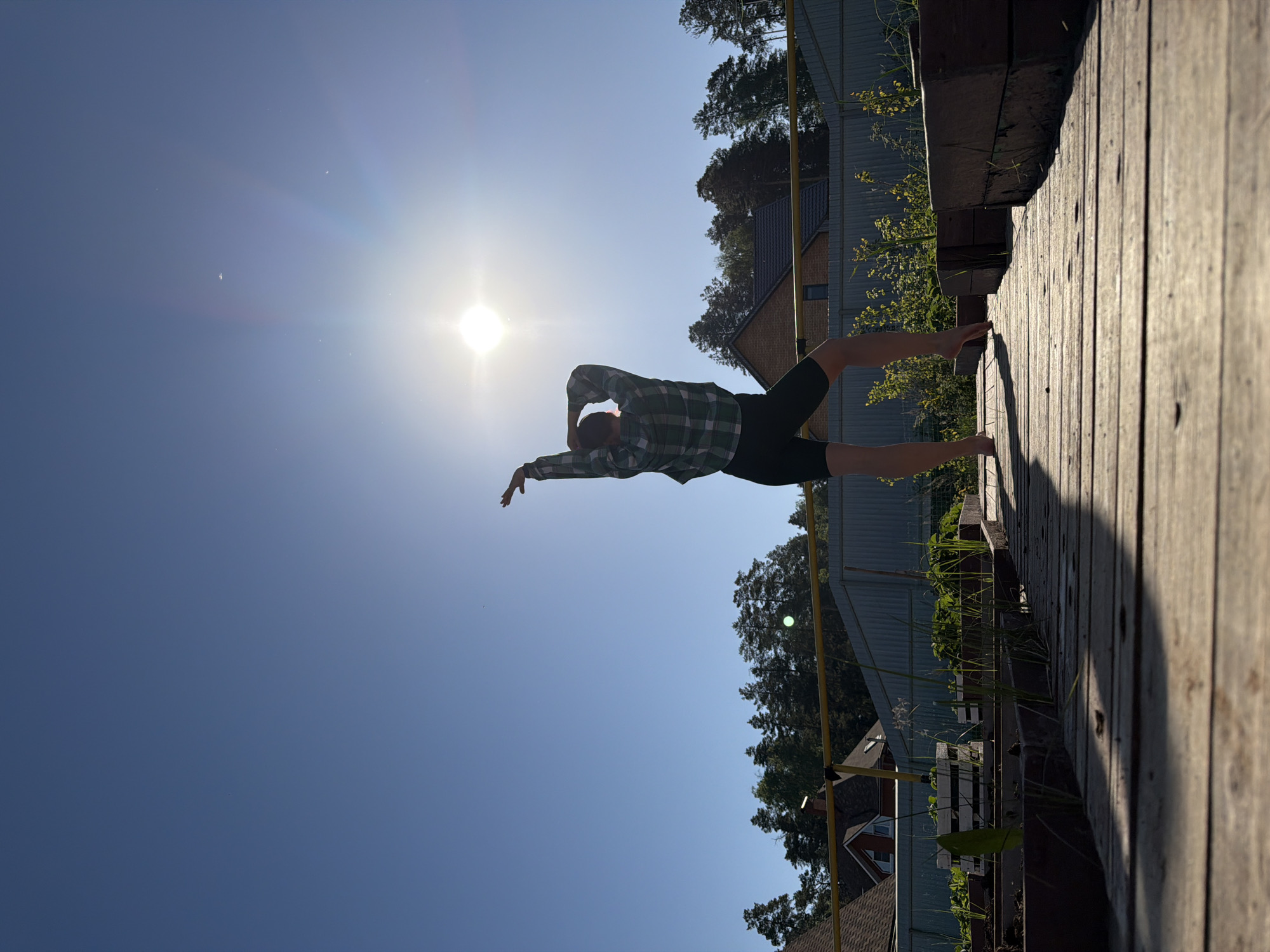 A person is standing on a wooden platform under a bright, clear sky with the sun visible. The person is wearing a green and black checkered shirt and black shorts. They are barefoot and have their arms outstretched, facing the sun. In the background are trees, a building with a brown roof, and part of a fence. The image was taken in Chaikovsky, Russia.